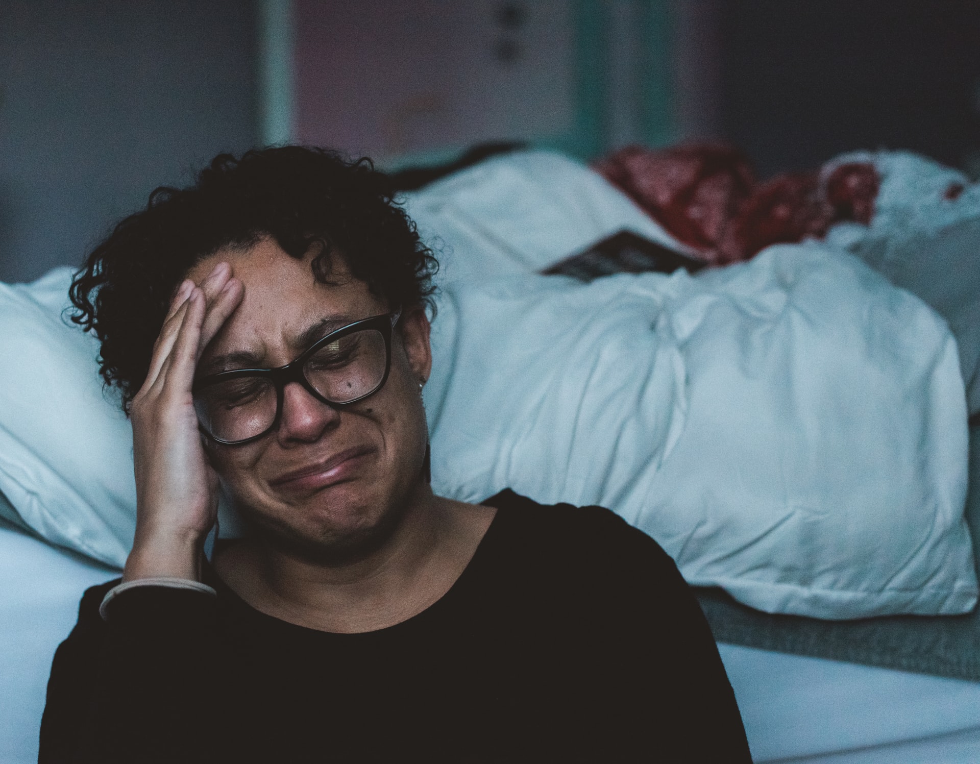 A woman holds her head and cries, sitting on the floor in front of an unmade bed.