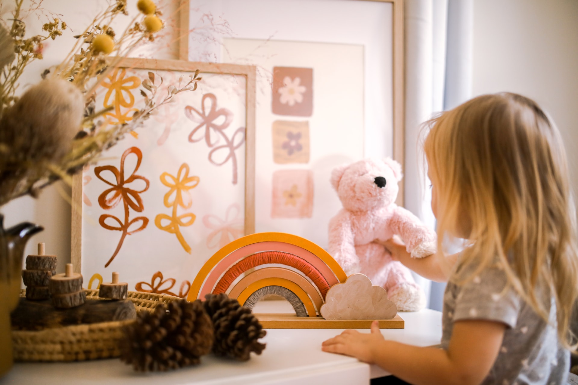 A little girl with blonde hair stands at a low shelf with artwork and a small teddy bear.