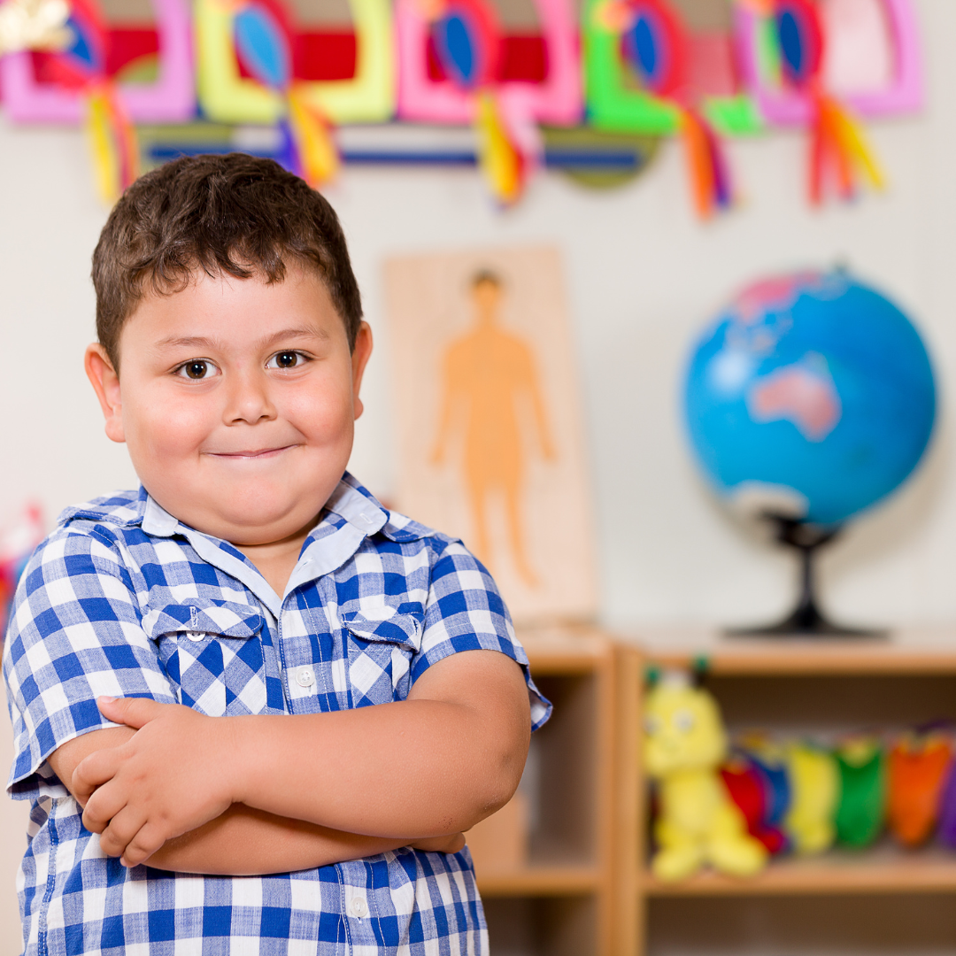 A six year old boy with short brown hair smiles at the camera.  He wears a blue and white checked shirt and has his arms crossed over his midsection.  He is standing in a classroom, with low bookshelves and a globe in the background.  There are colorful pictures which appear to be children's artwork on the walls in the background.