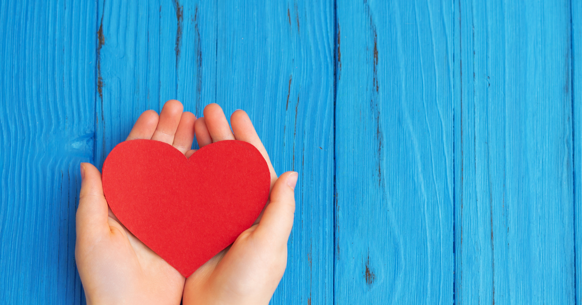 A child's hands hold a red paper heart over a blue painted wooden background.