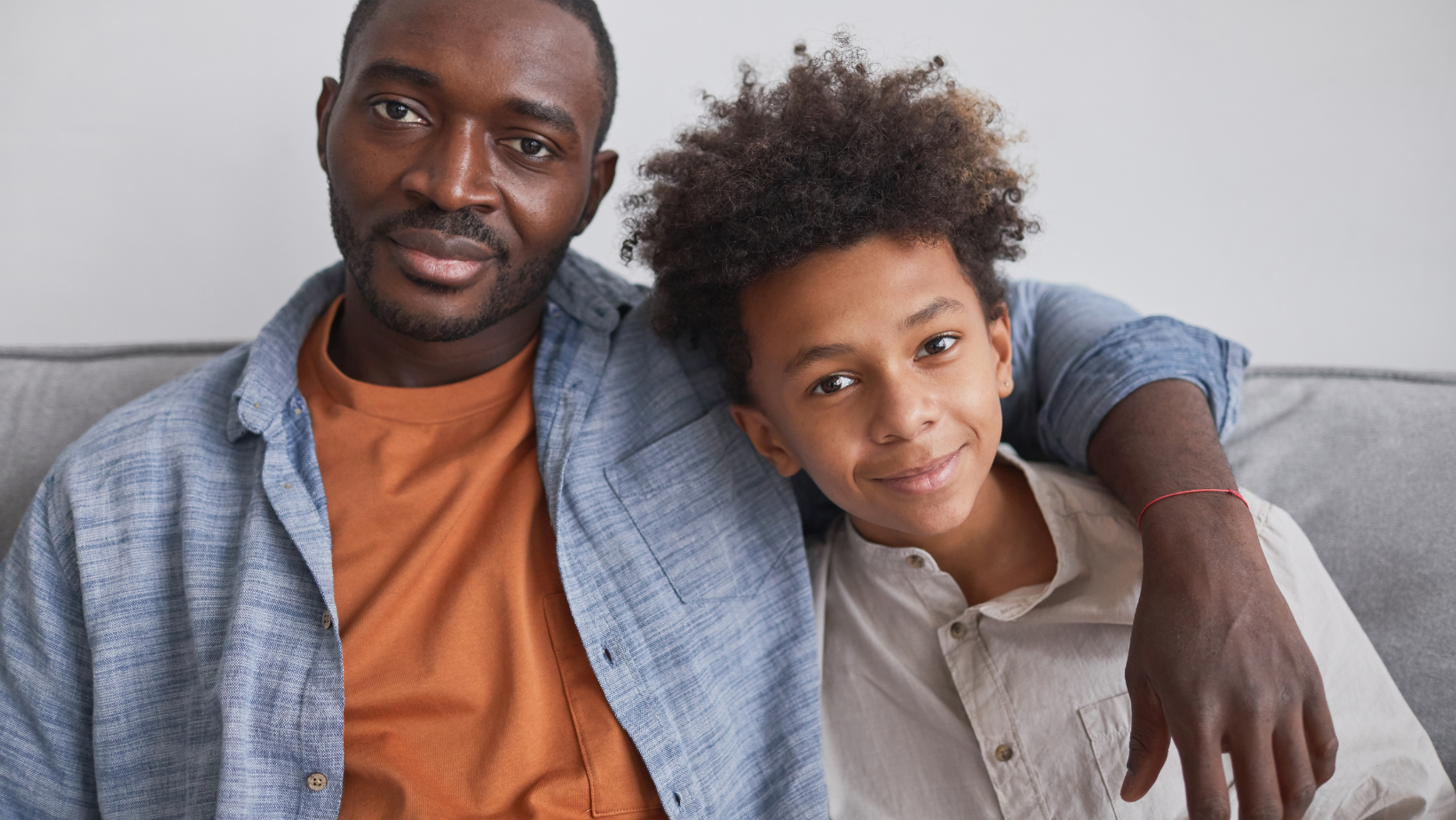 A Black male sits on a couch with his arm around a biracial teen boy, who is smiling.