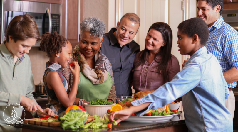 A multi-racial, multi-generational group of people gather around a kitchen island.  They are chopping vegetables and have a holiday turkey ready.  Most people are laughing and engaged, but one teenage boy is cutting vegetables and appears disconnected from the others.