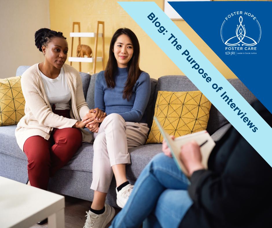 A Black woman and an Asian woman sit on a couch holding hands, looking at an interviewer. The interviewer in is the foreground and the back of their left shoulder and side are seen. The interviewer appears to be a woman, wearing jeans and a sweater. She holds a notepad and pen.