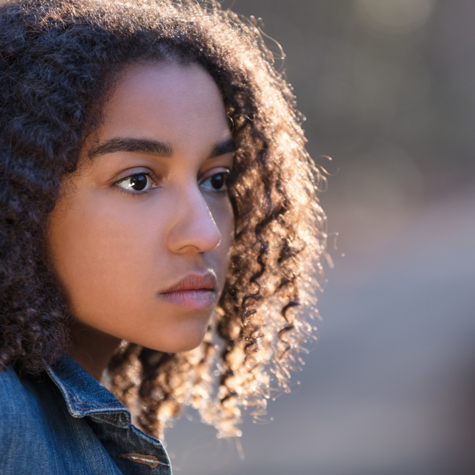 A teenage, Afro-Latina girl stares into the distance.  She has curly, dark brown hair and wears a jean jacket.