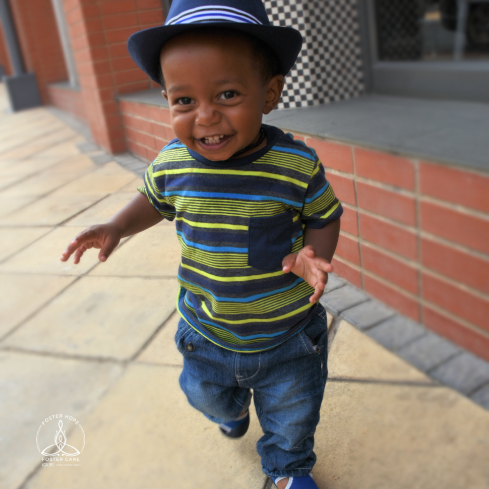 A toddler boy in a striped shirt and a panama-style hat smiles at the camera.