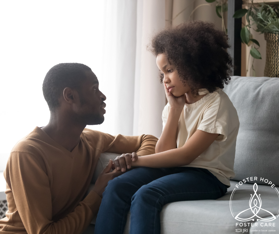 A foster father kneels in a supportive posture beside an eight year old child who appears upset.