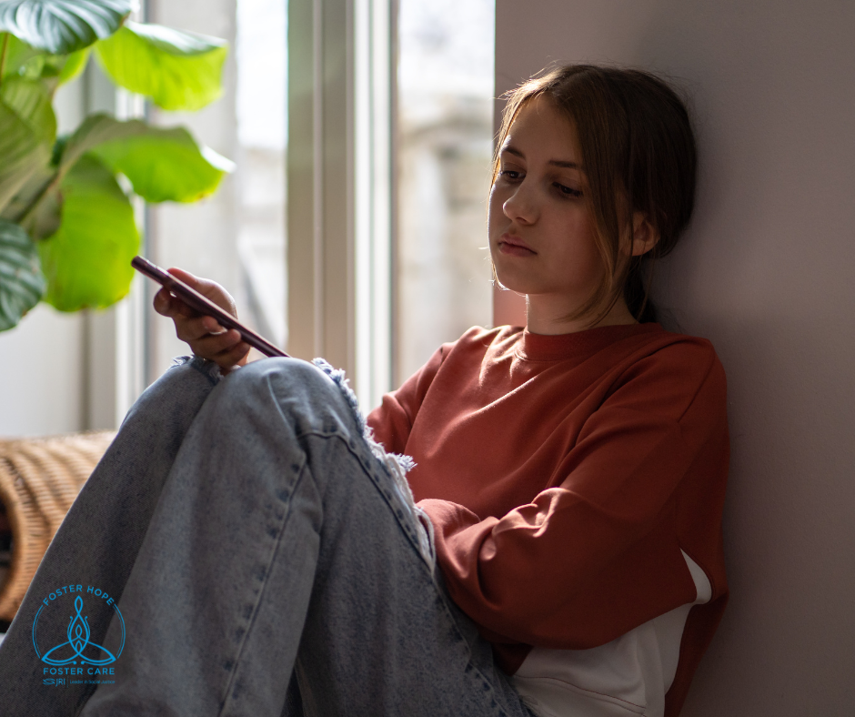 A teenage girl slouches, seated on the floor with her back braced against a wall.  She holds a cell phone in her hand.  She has a distressed expression on her face.