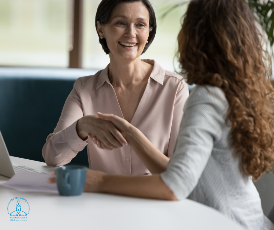 Two women sit at a table with a blue cup of coffee and shake hands to greet one another warmly.