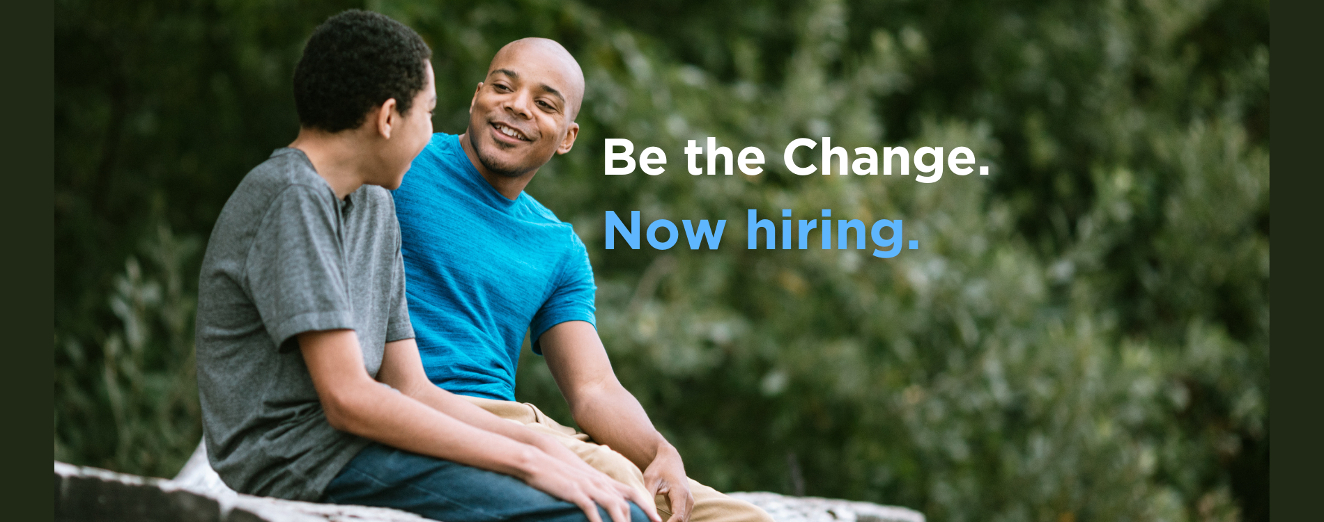 A photo of young male of color with and adult male of color sitting on a rock talking.  Text says Be The Change.  Now Hiring.