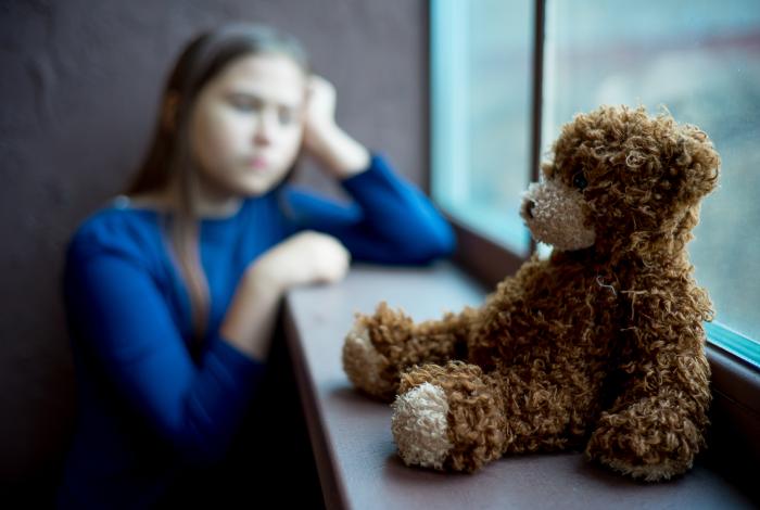 young girl looking out window with teddy bear in front of her