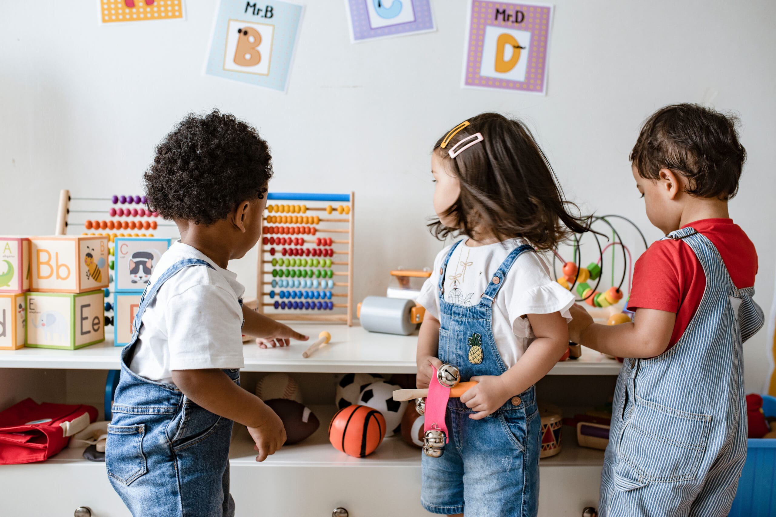 Three children in overalls playing with toys in a room with educational decorations.
