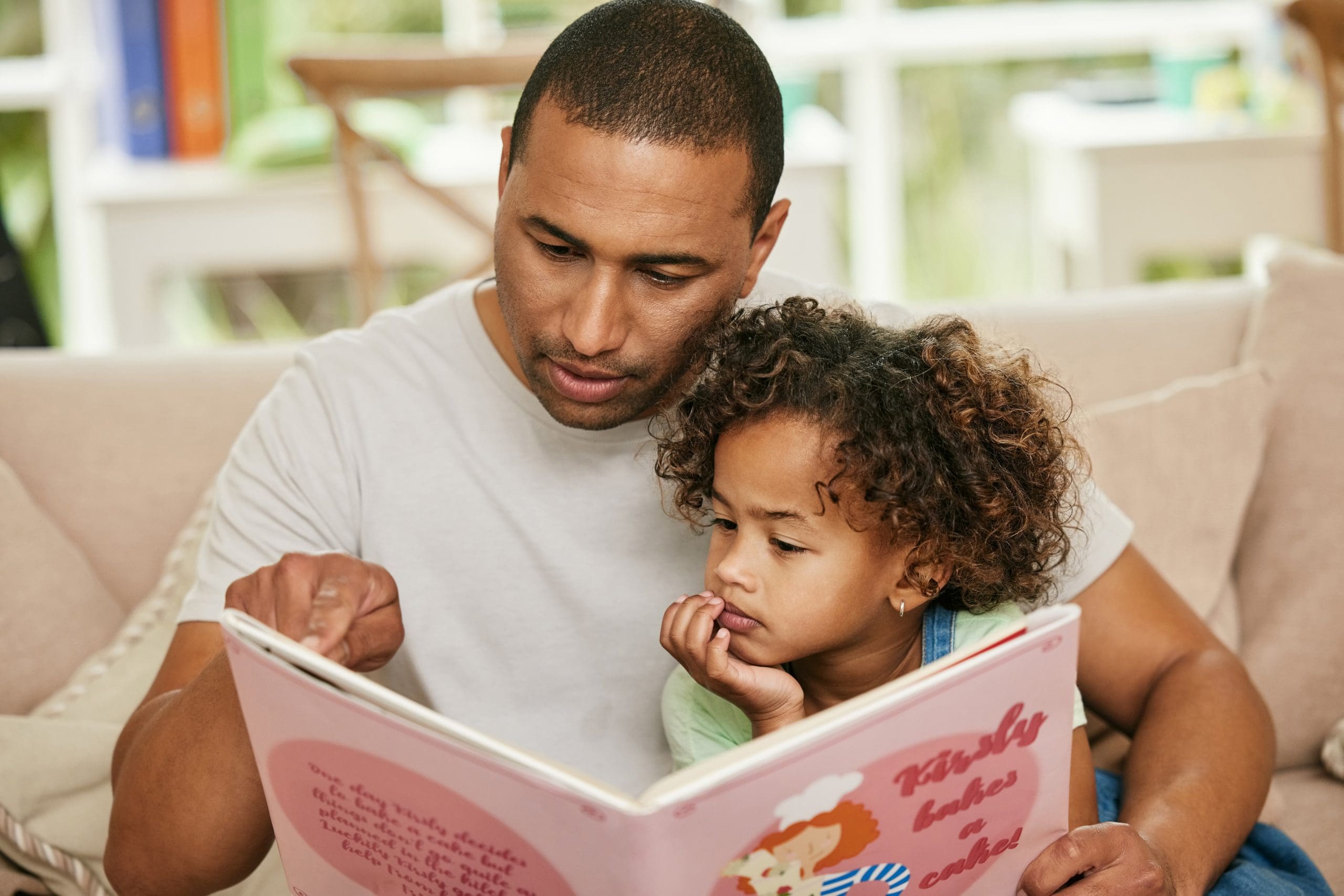 Father reading a book to a young girl with curly hair on a sofa.