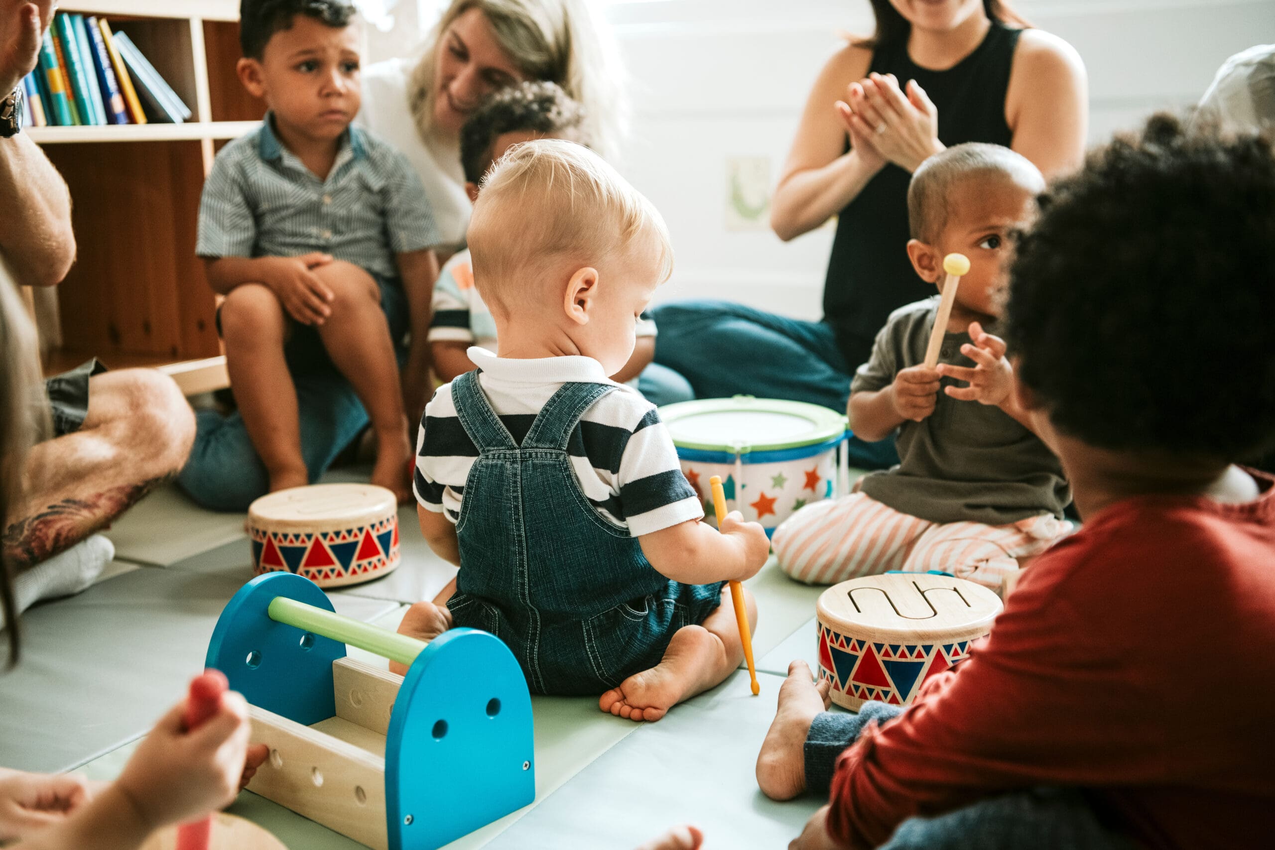 Children and adults sitting on the floor during a group activity, with drums and musical toys around.