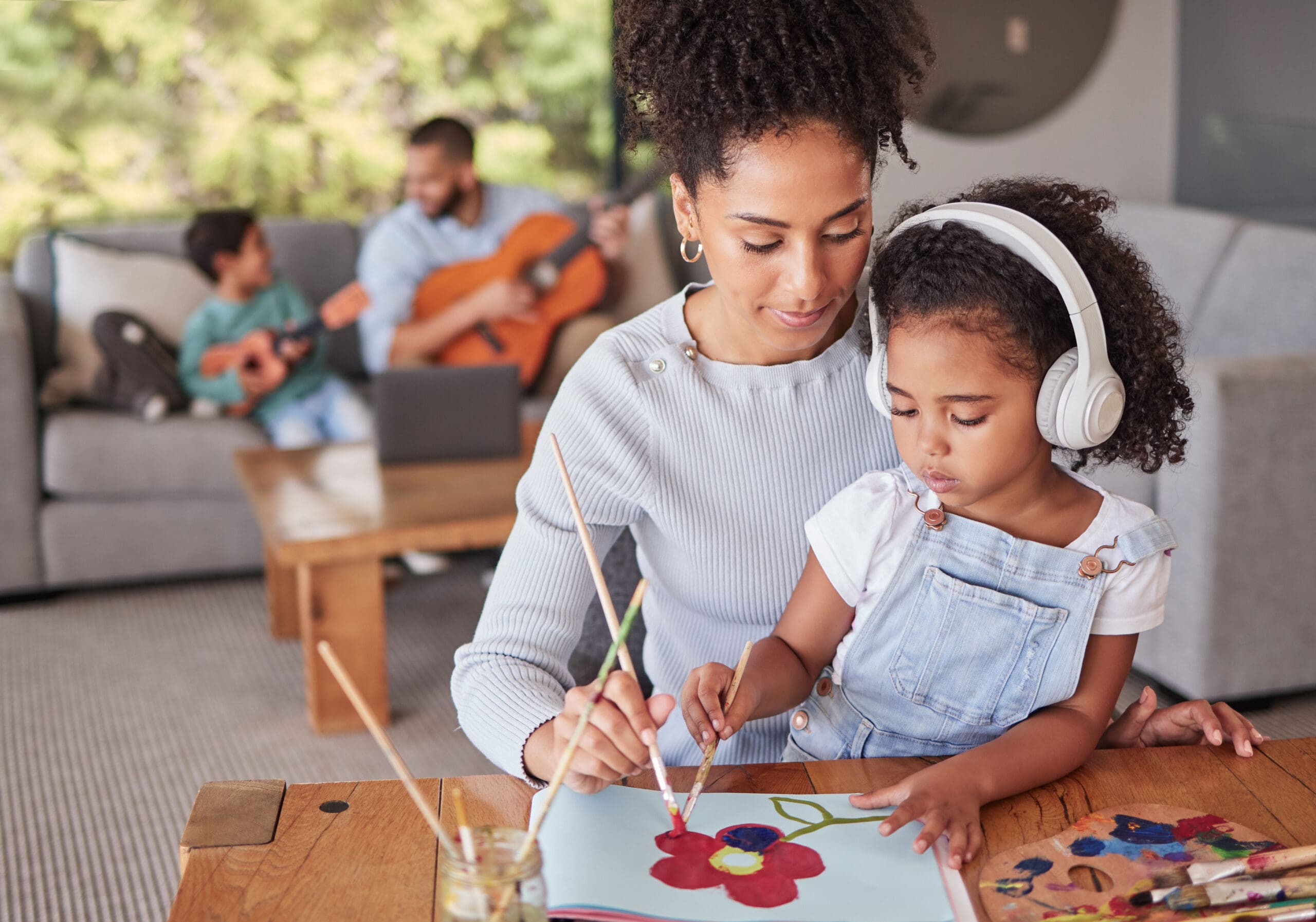 Mother helping daughter paint a flower while father and son play guitar in the background.