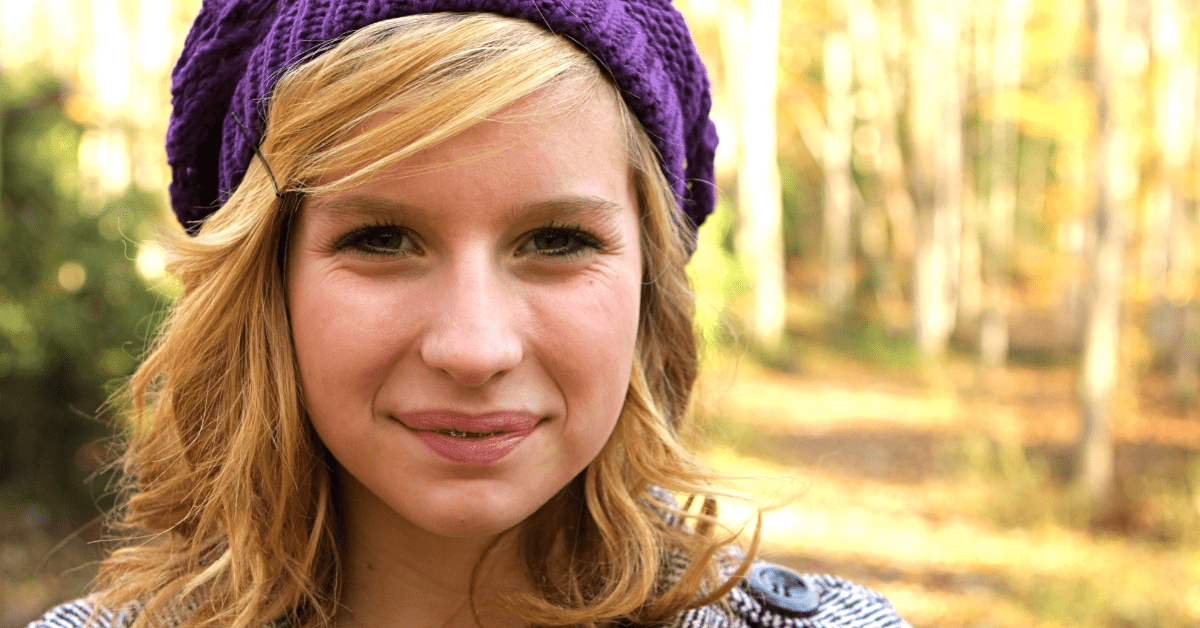 Close-up of a smiling person with blonde hair and purple hat in a wooded area