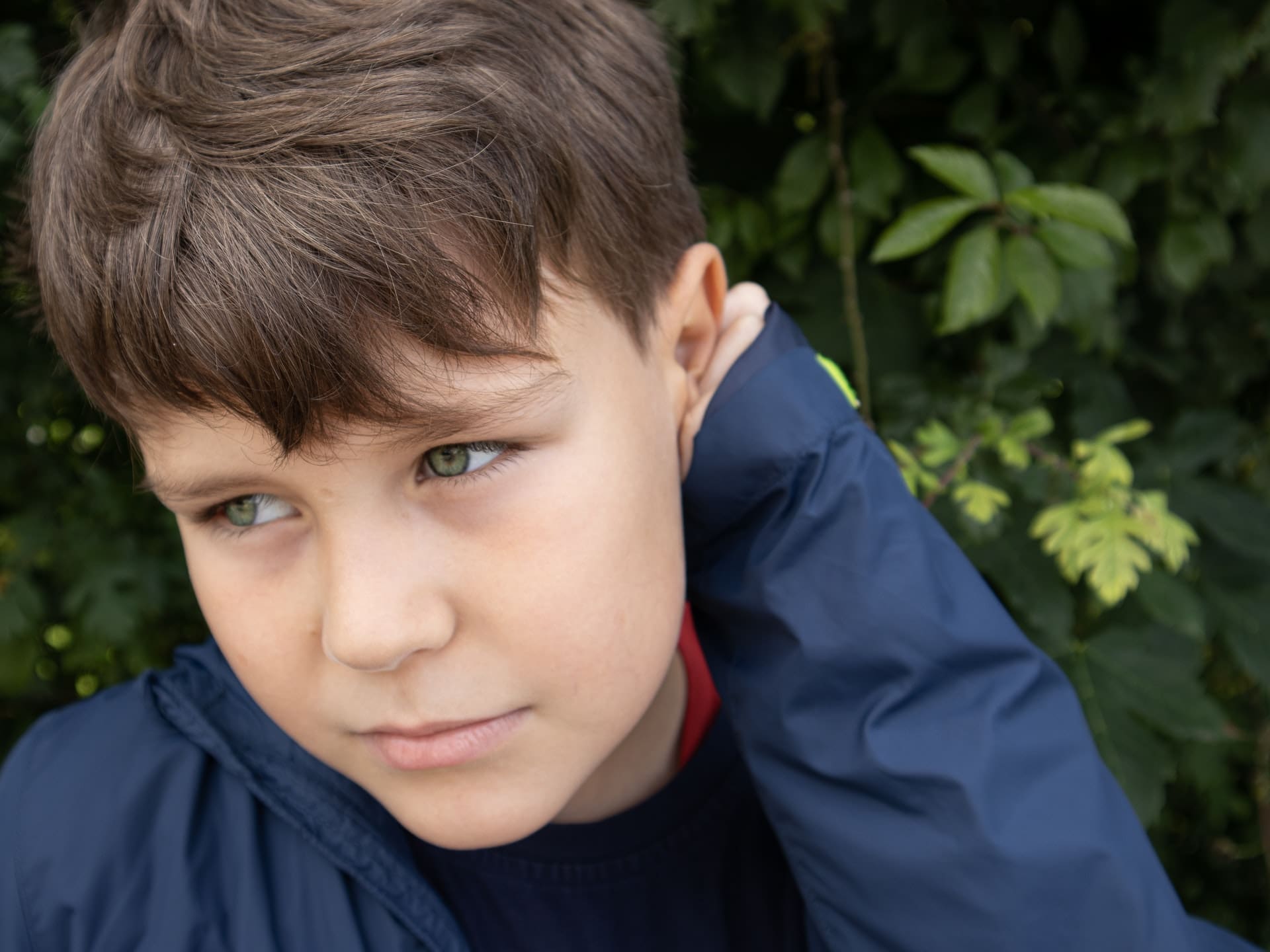 Close-up of a child with short brown hair and green eyes, looking to the side, in front of green foliage.
