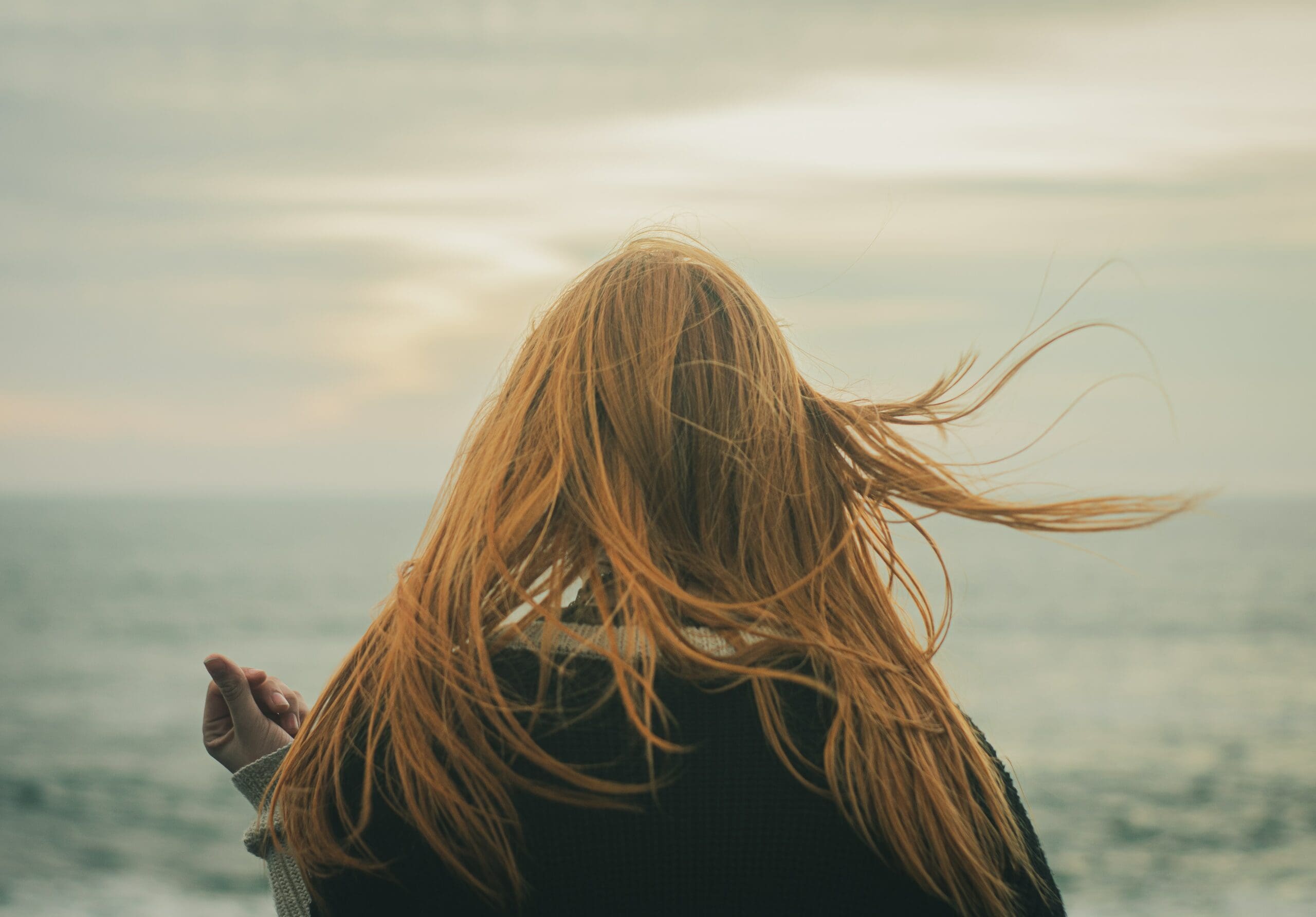 Person with long red hair facing water, wind blowing hair.