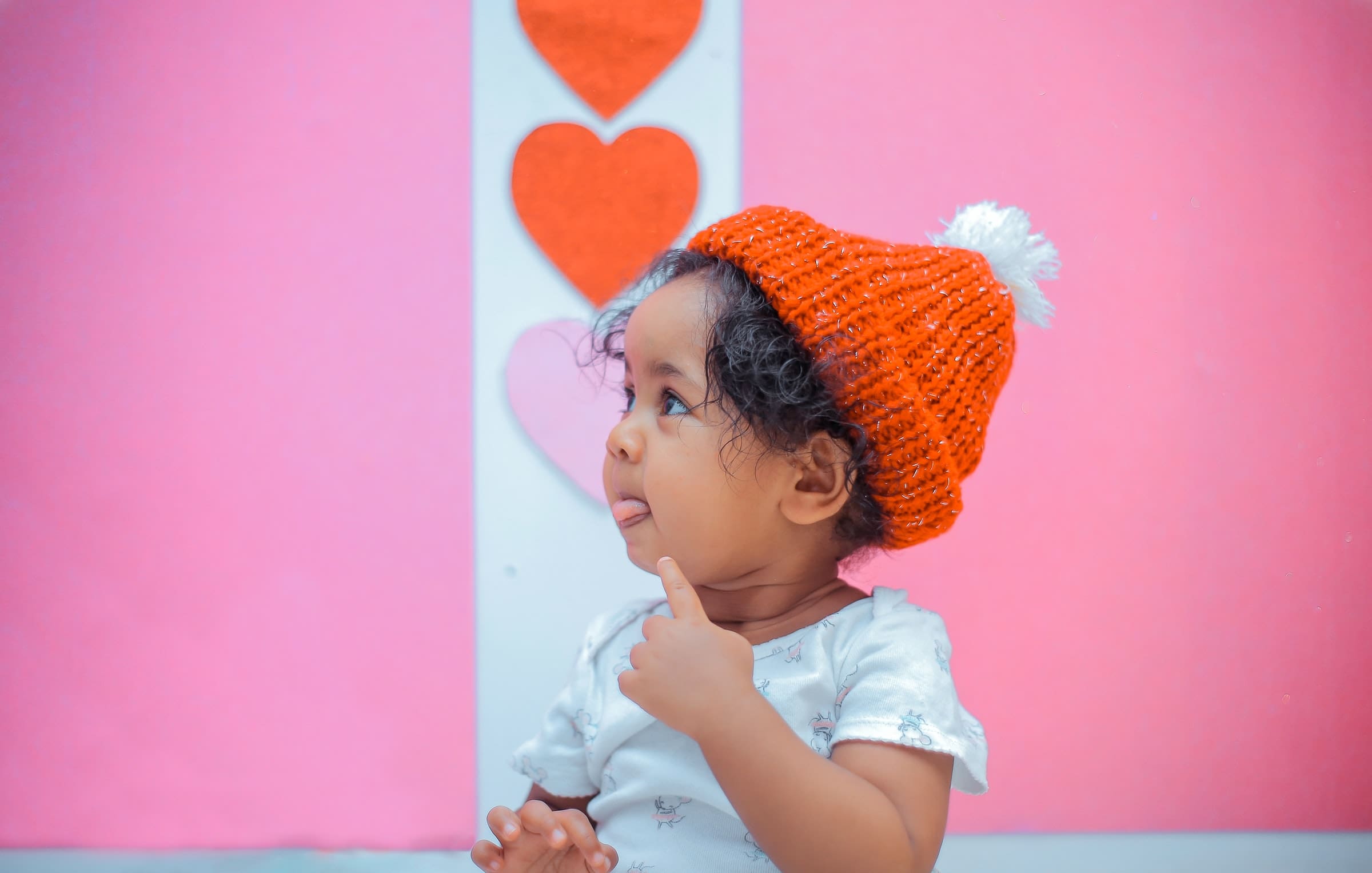 Child wearing a red knit hat with a white pom-pom, pink background with hearts
