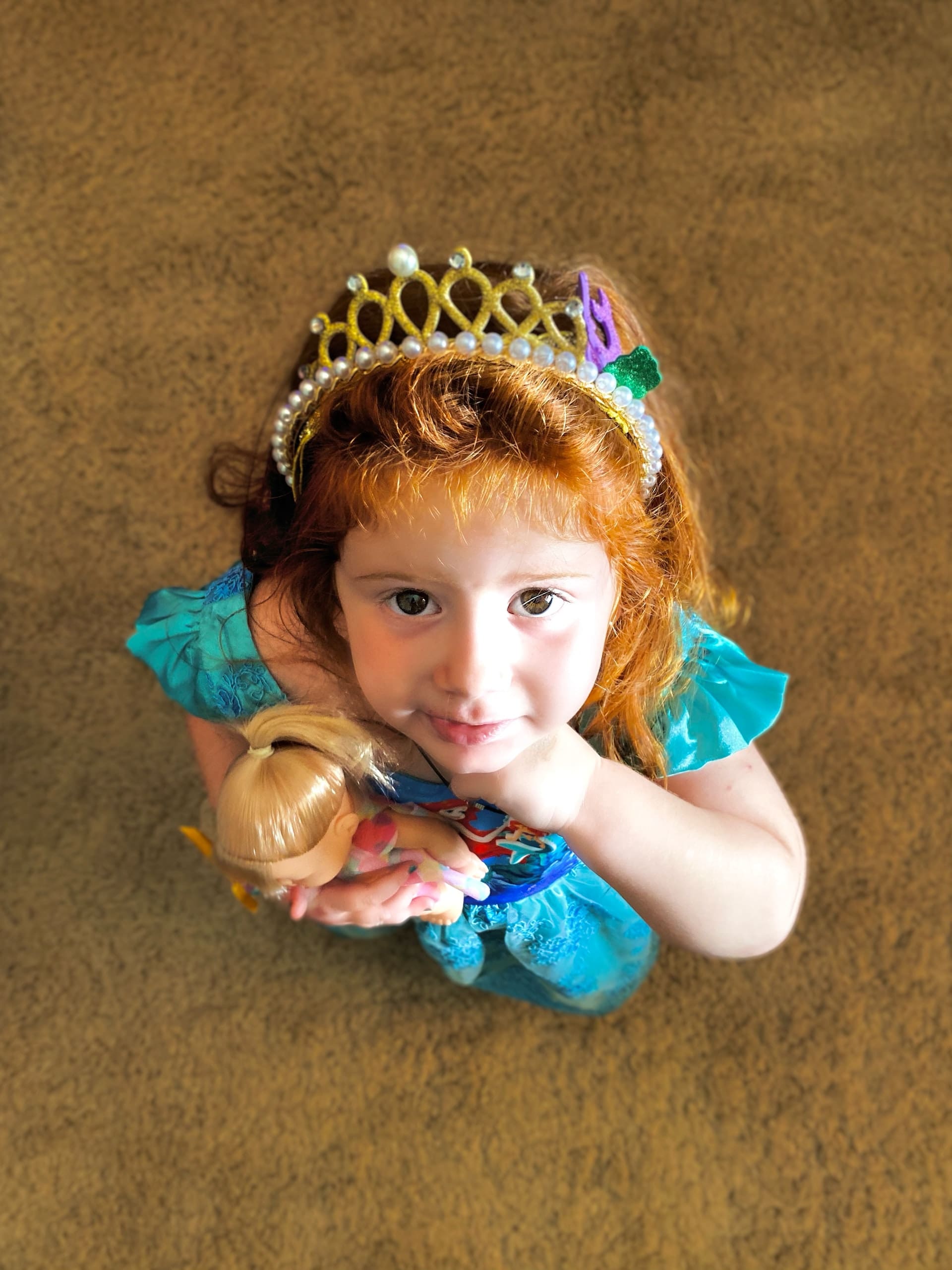 Child with red hair wearing a blue princess dress and crown, holding a doll.