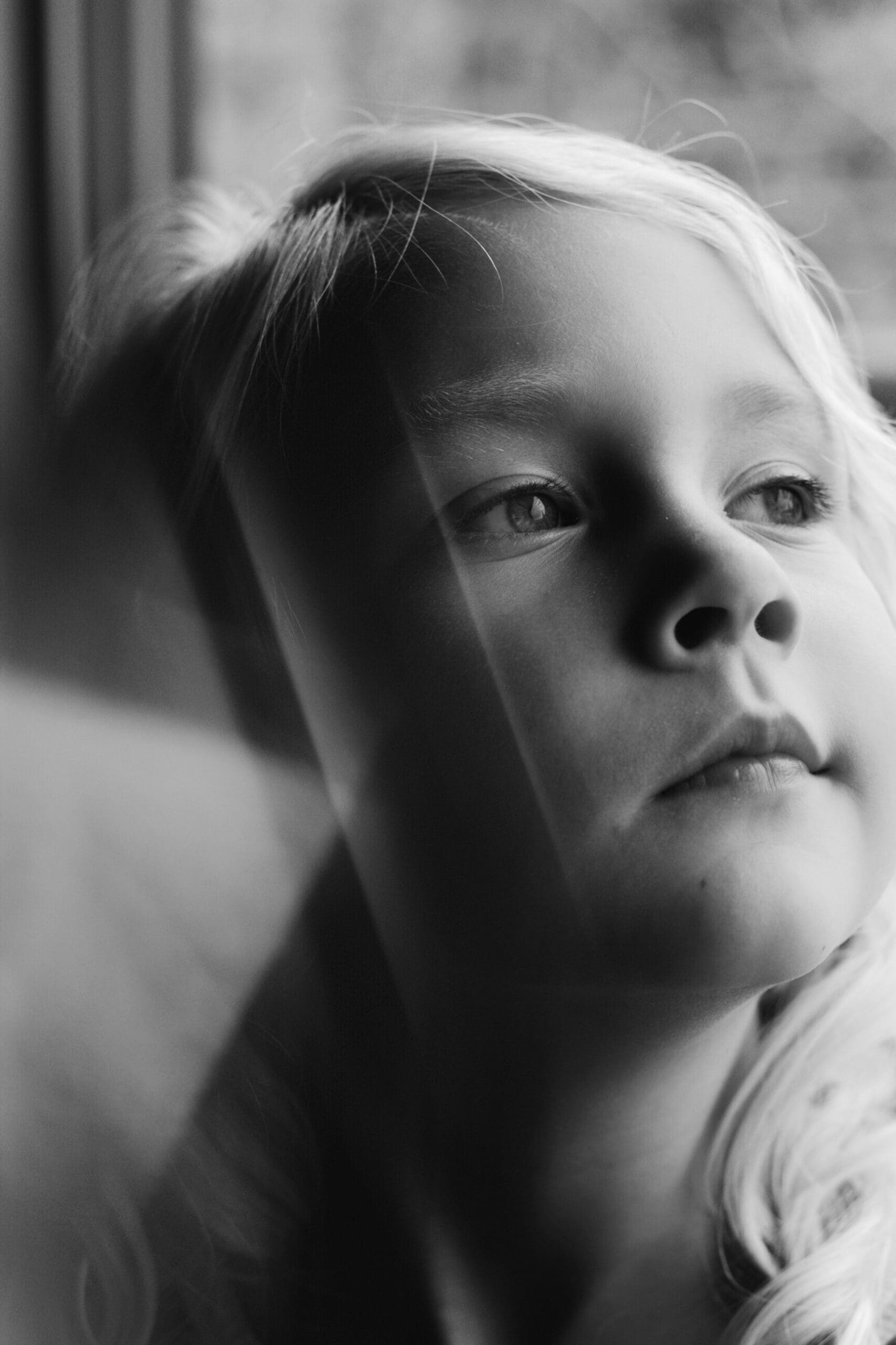 Child looking out a window with light reflecting on their face in black and white.