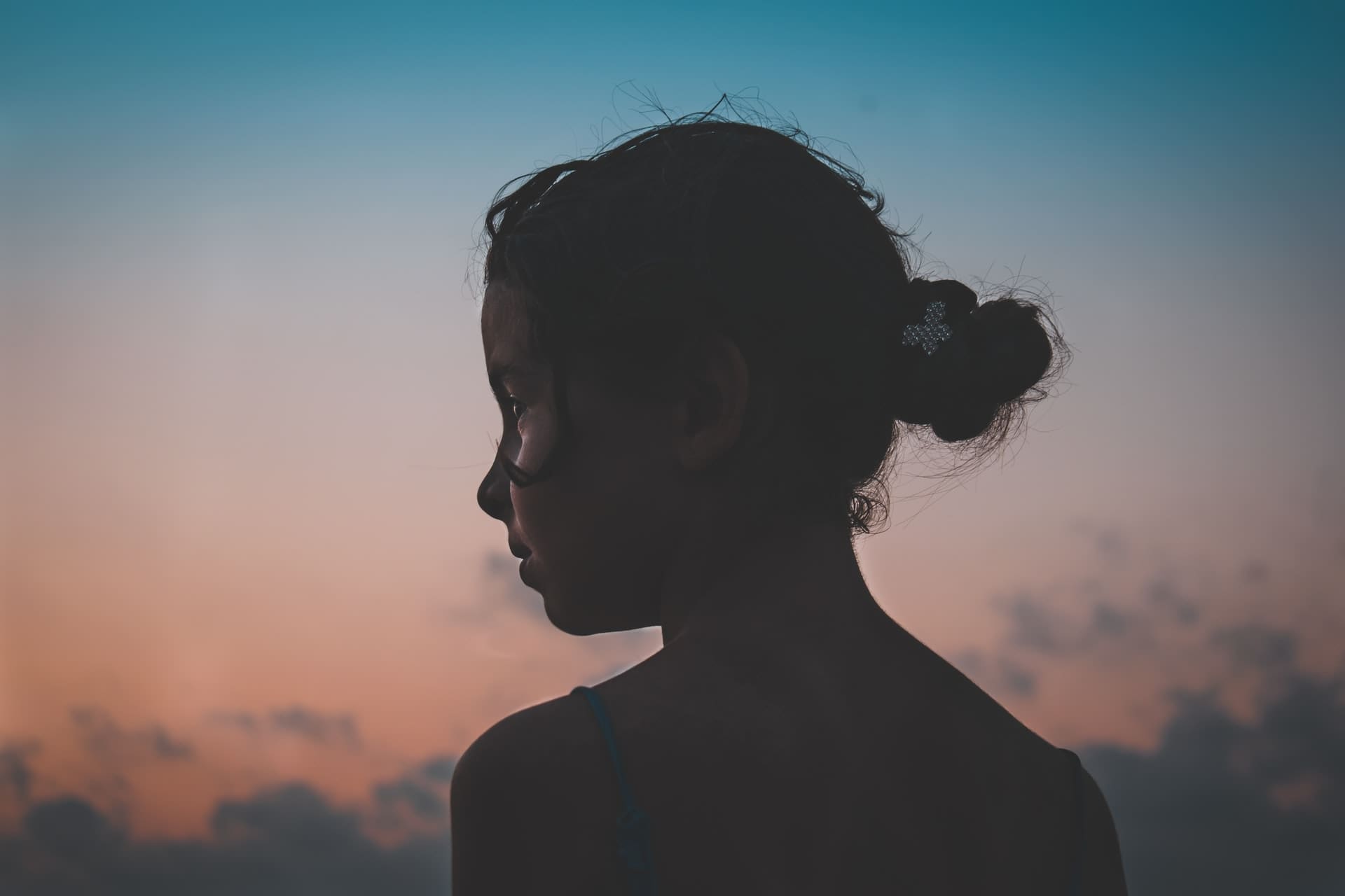 Silhouette of a child with hair in a bun, looking away, against a sunset sky.
