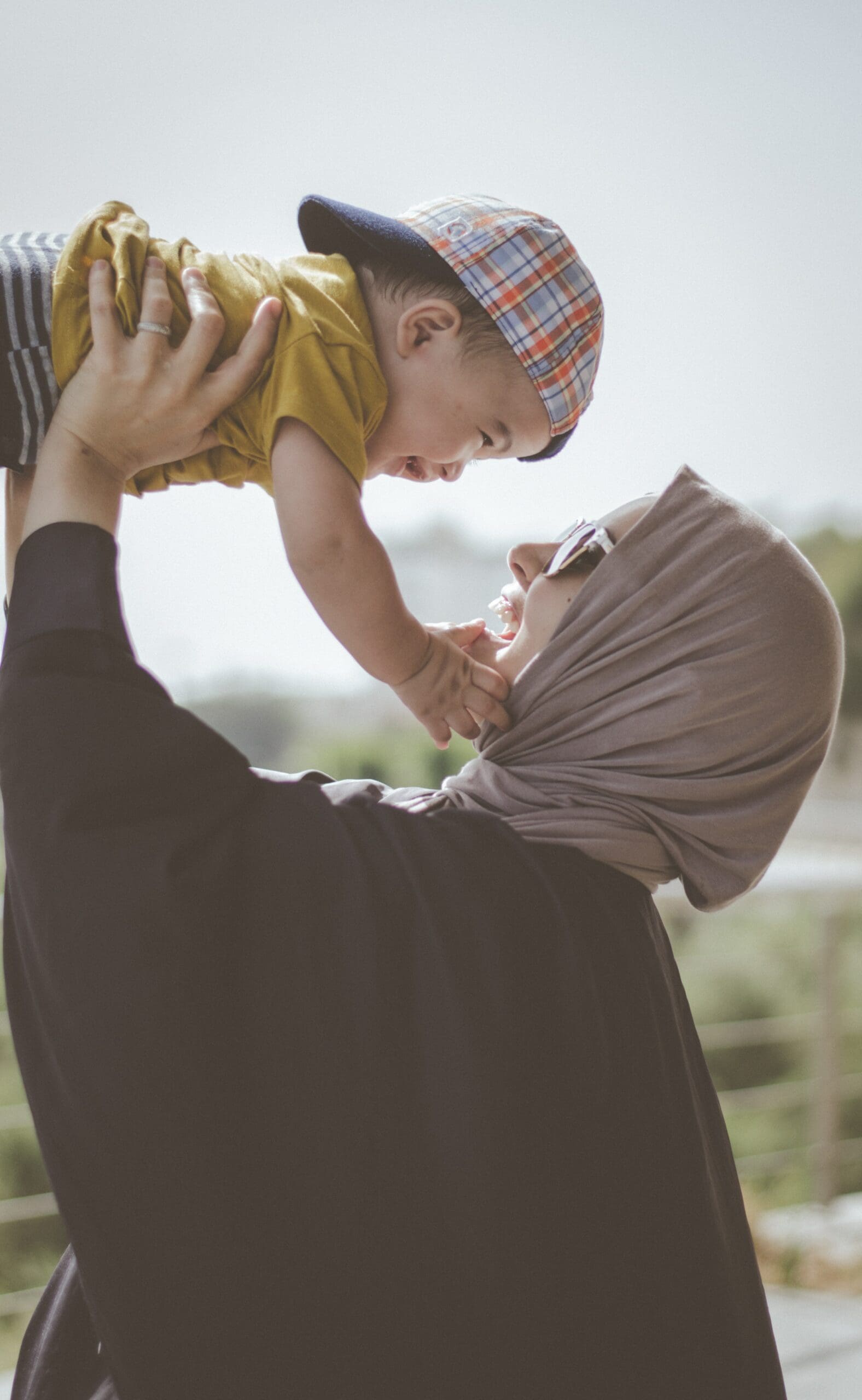 Person wearing hijab holding a child up, child's face near their face, outdoors.