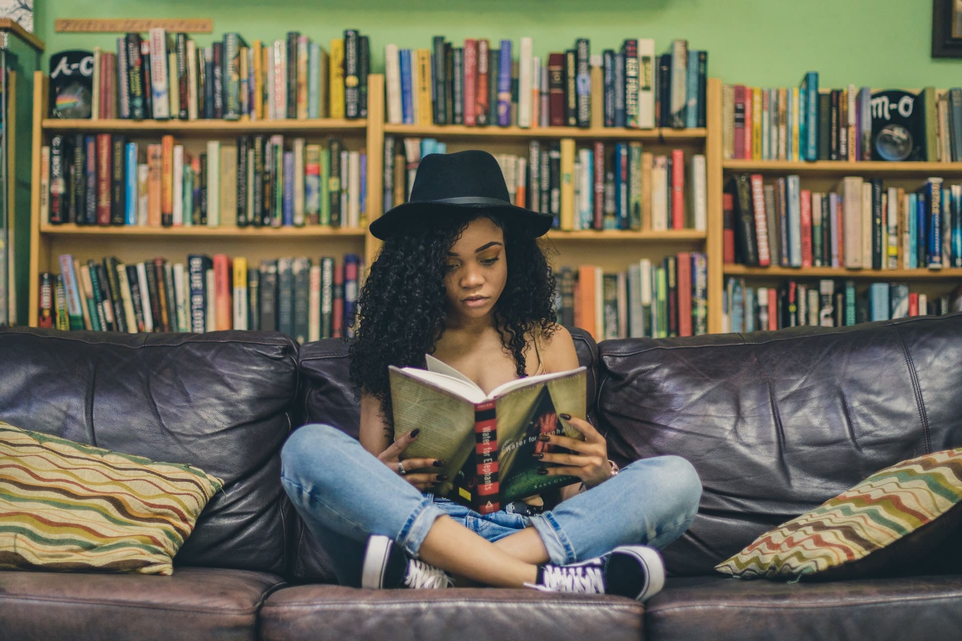 Person with curly hair and hat reading a book on a couch with a bookshelf in background.