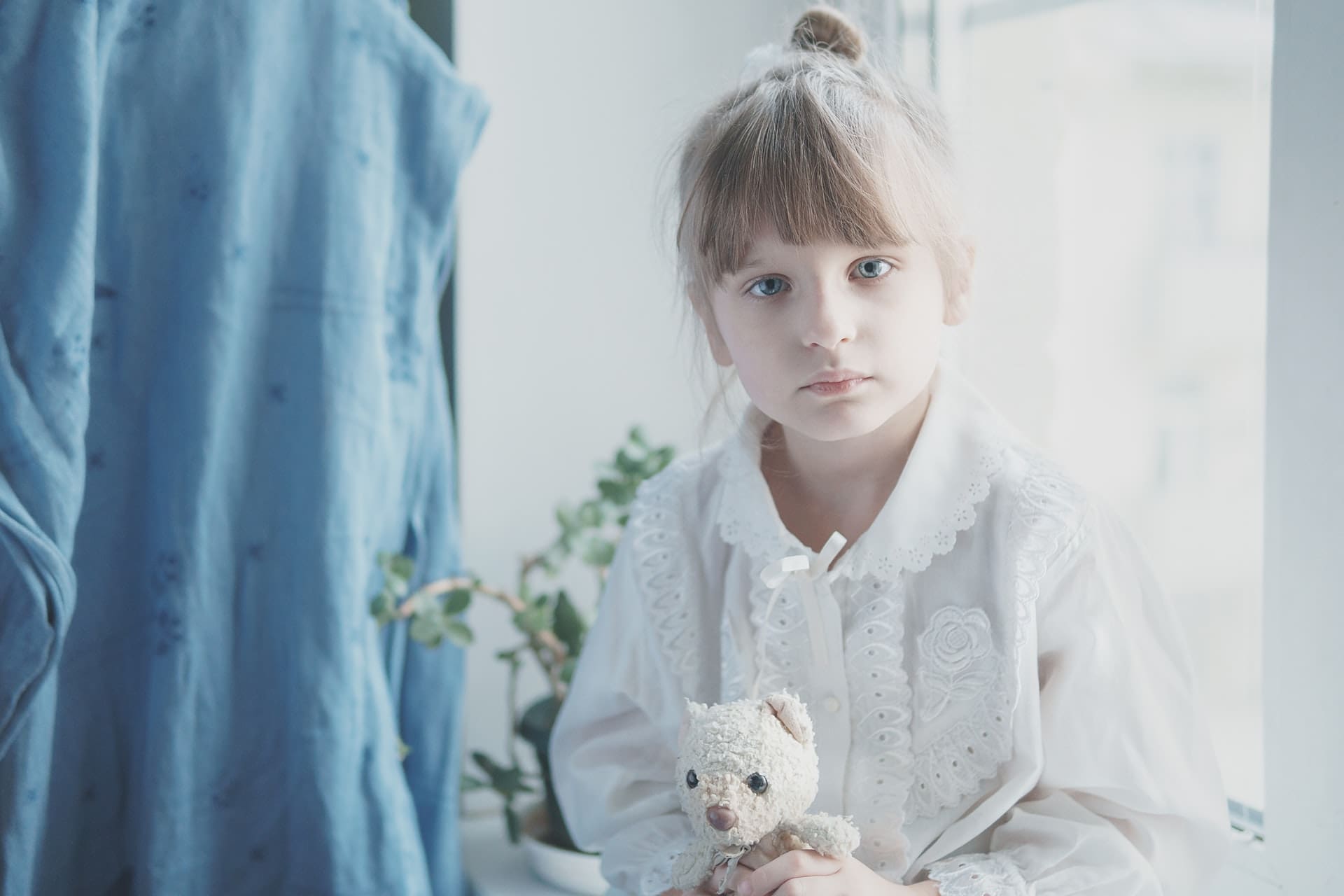 Child with light hair and blue eyes holding a teddy bear near a window.