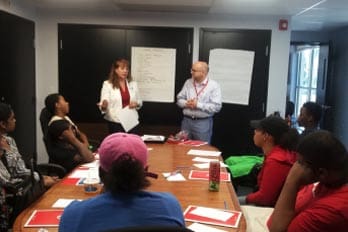 Two presenters standing at the front of a room with a group seated around a table.