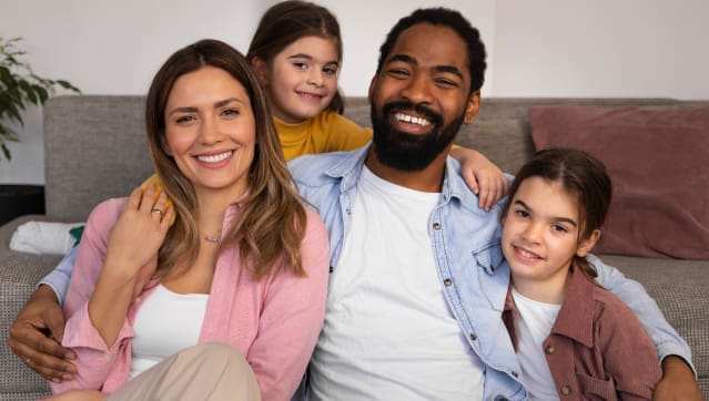 Family sits together on a couch smiling, with two children.
