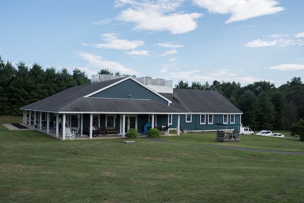 Large blue building with a porch and white chairs, surrounded by grass and trees.