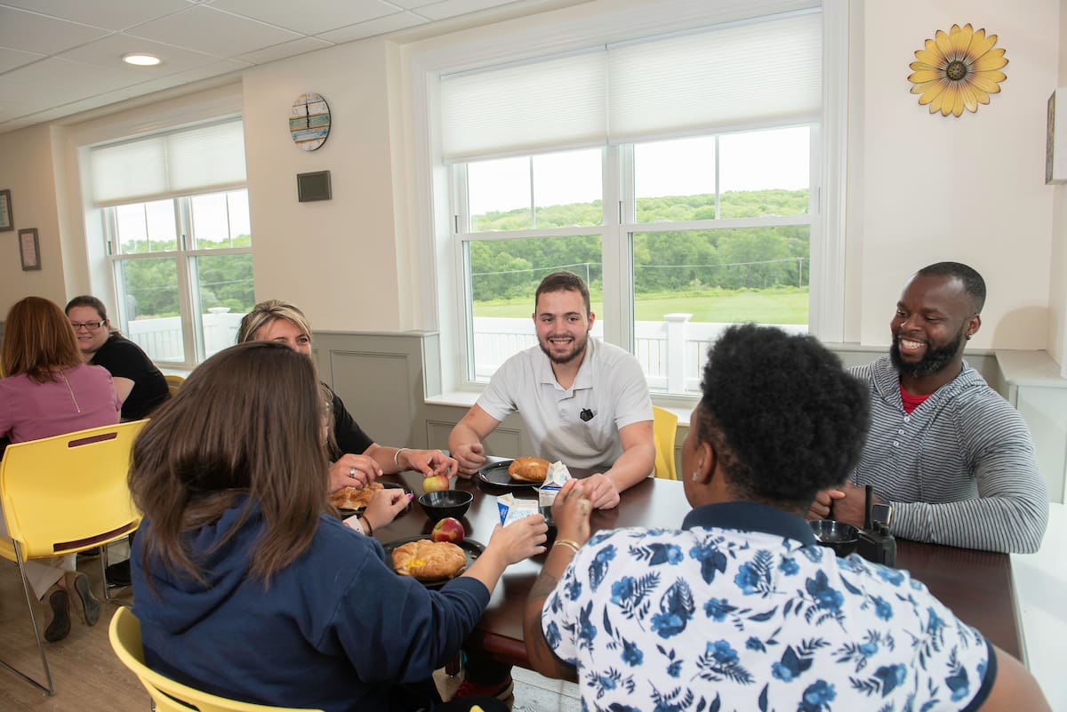 Group of people smiling and eating together at a table in a bright room with large windows.