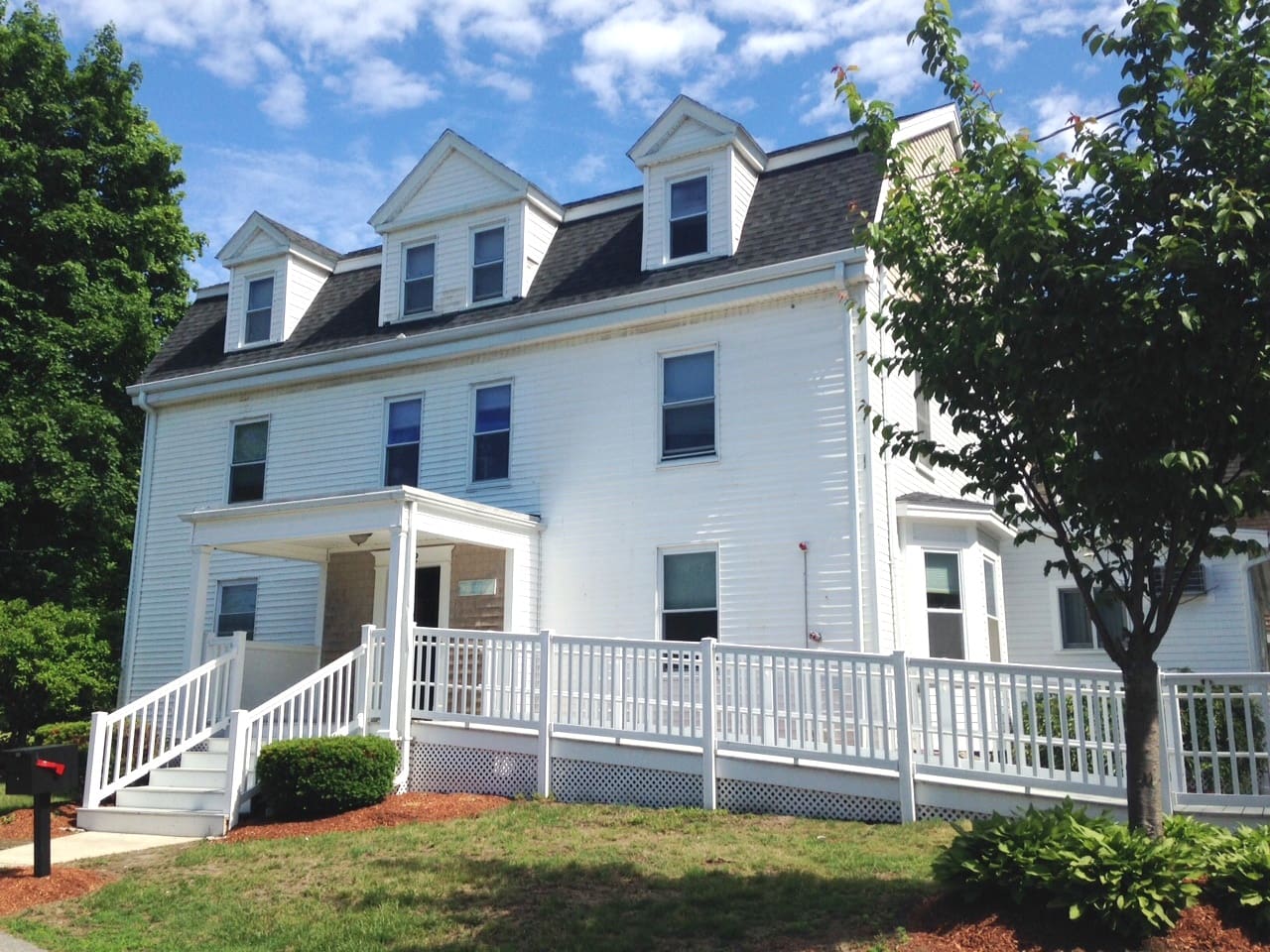 White multi-story house with a porch, surrounded by greenery and a blue sky