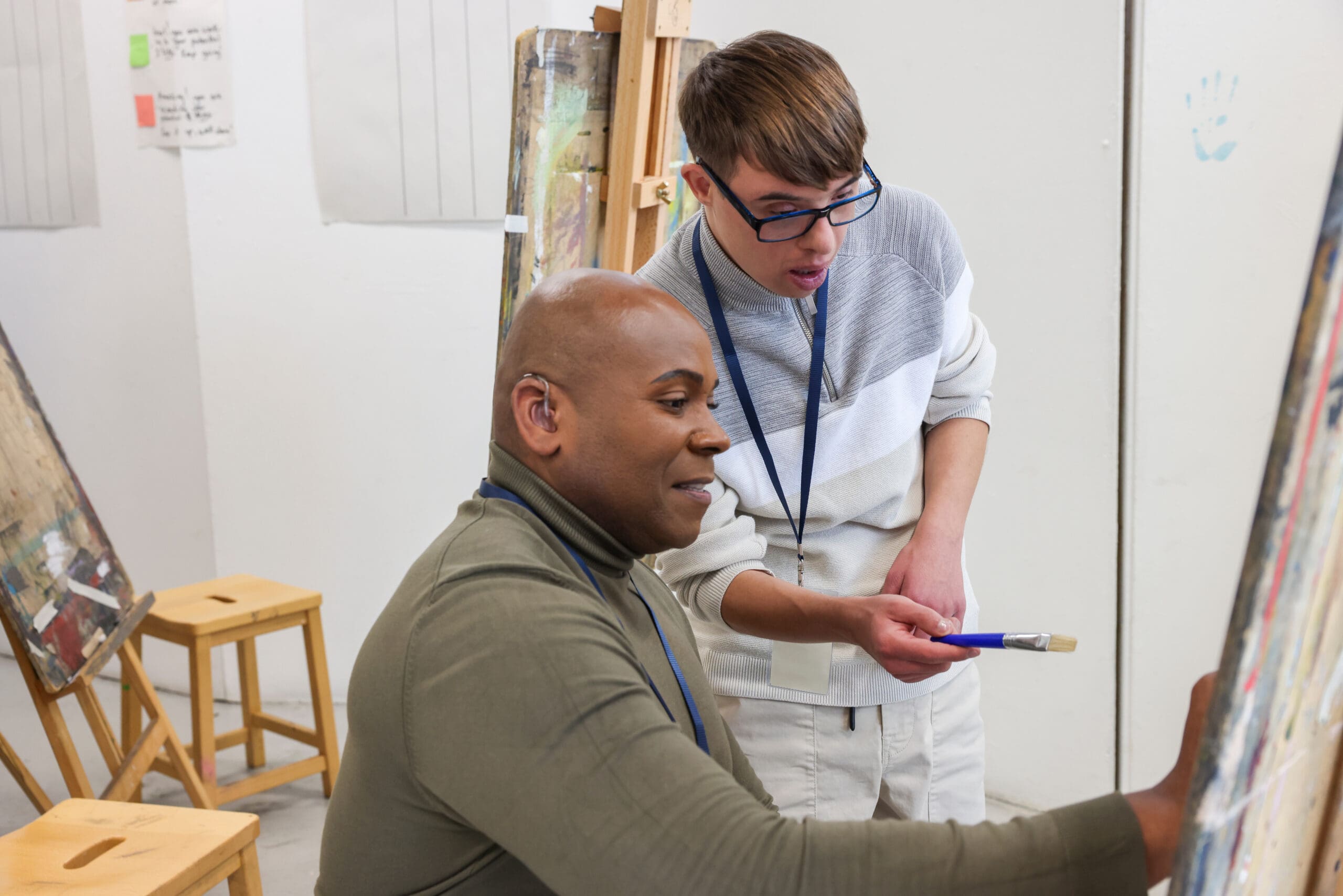 Two men, one seated and painting, the other standing and discussing, in an art studio.