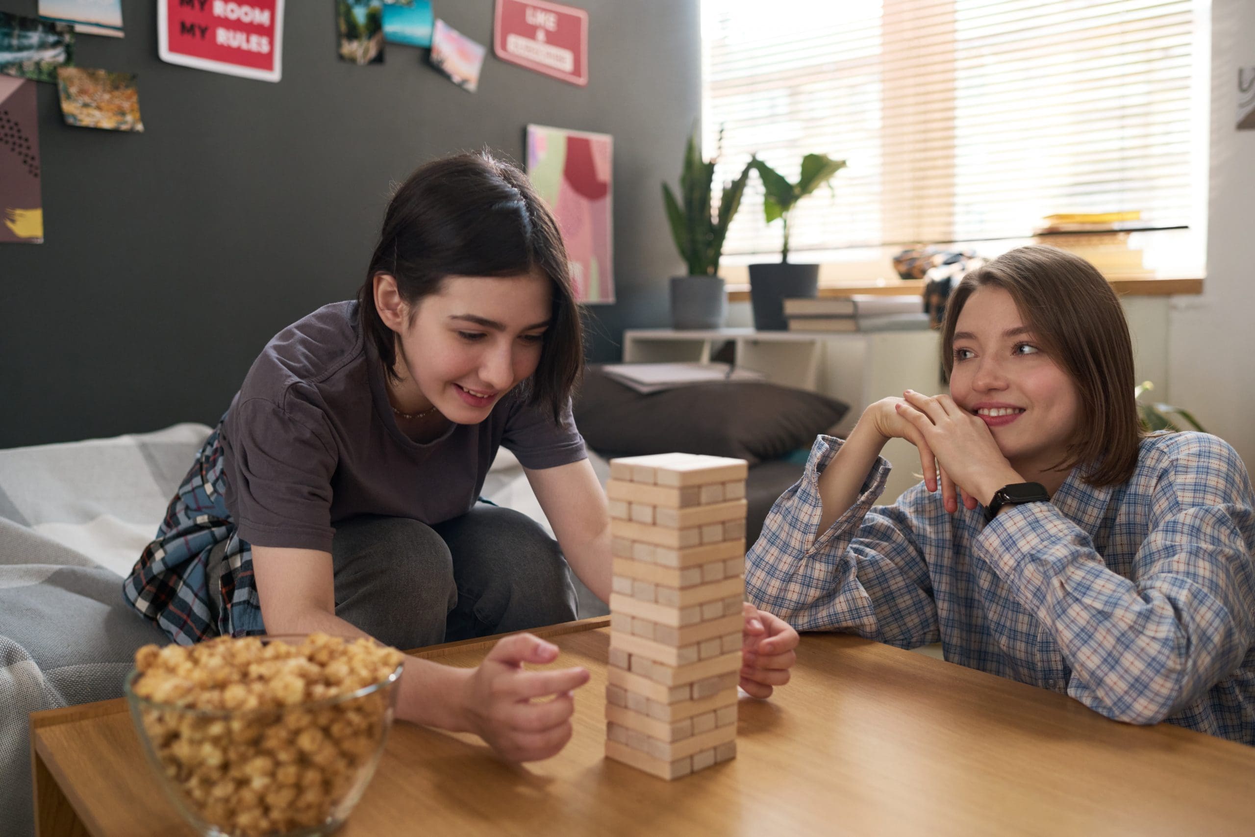 Two teenagers playing Jenga at a wooden table, with a bowl of popcorn nearby.