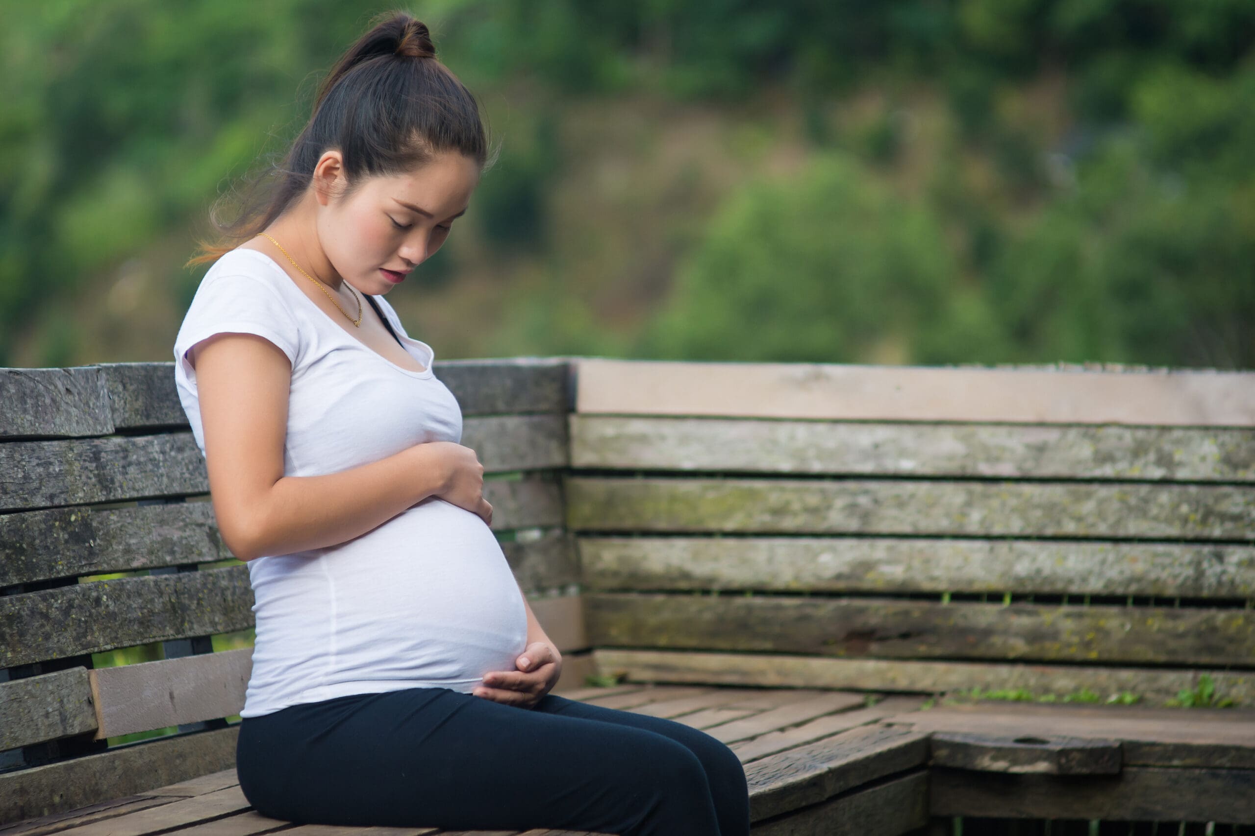 Pregnant woman sitting on a wooden bench outdoors, looking down at her belly.