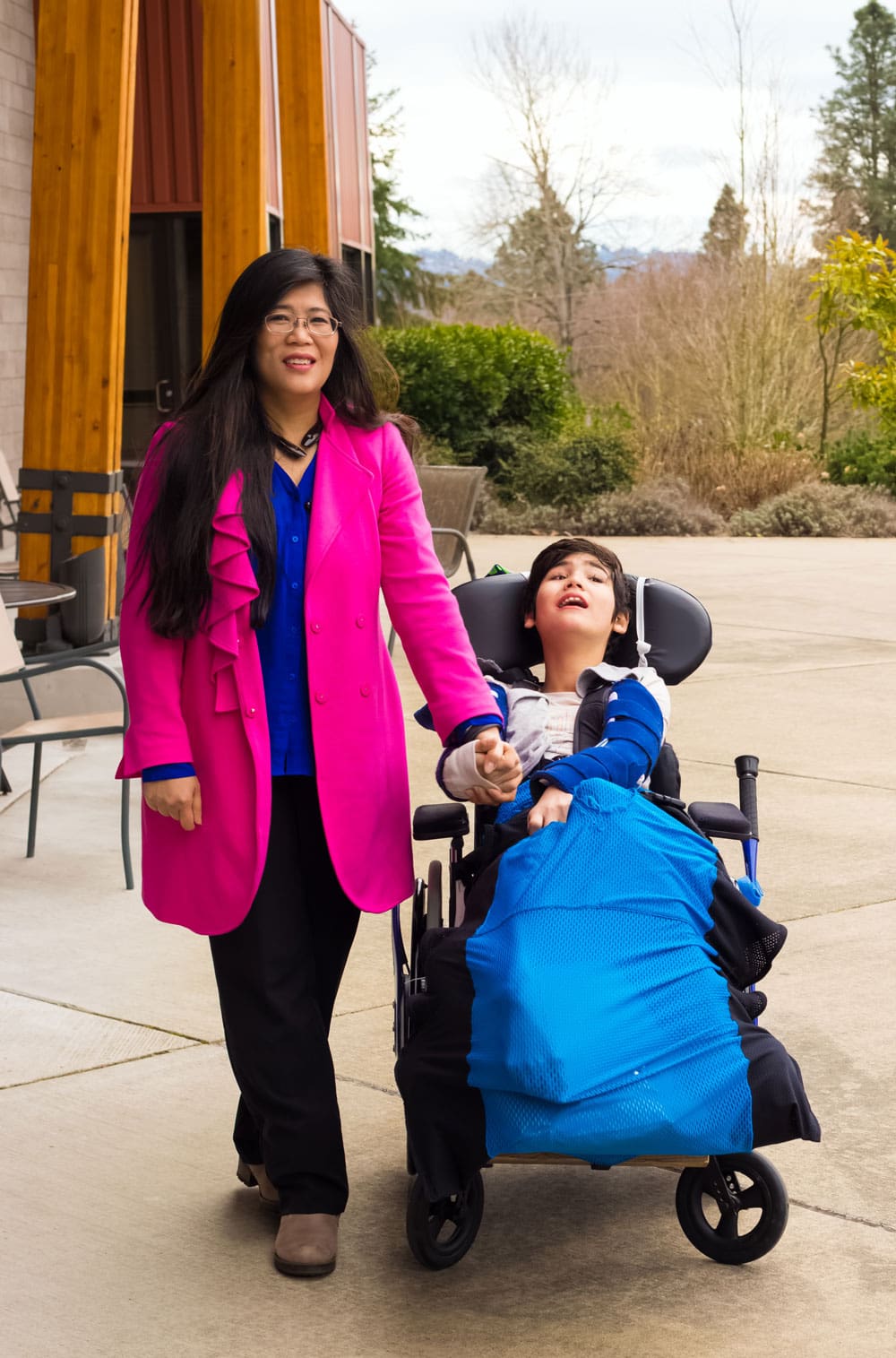 Woman and a boy in a wheelchair outside holding hands.