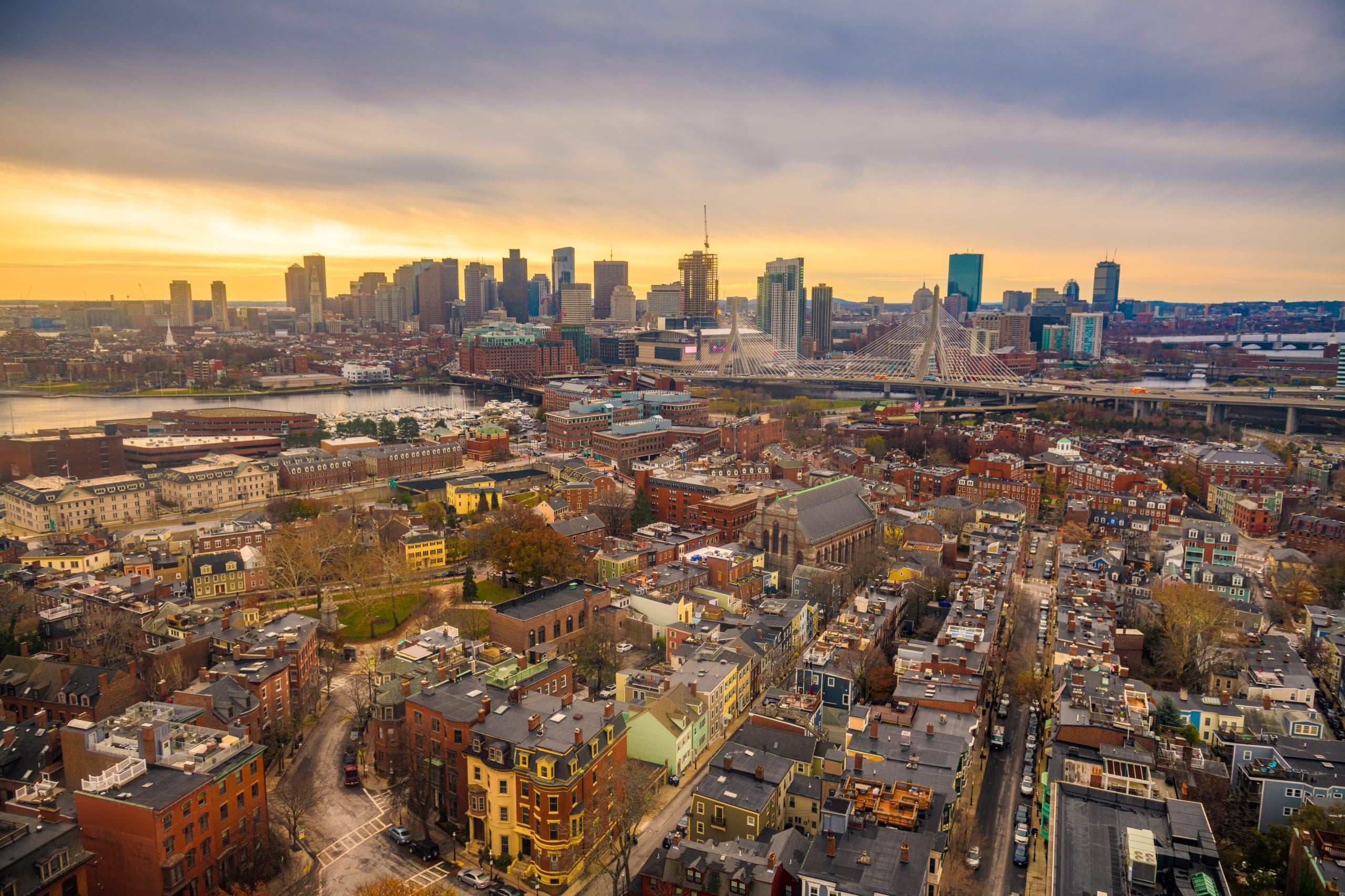 Boston skyline with high-rise buildings and bridge over water at sunset.