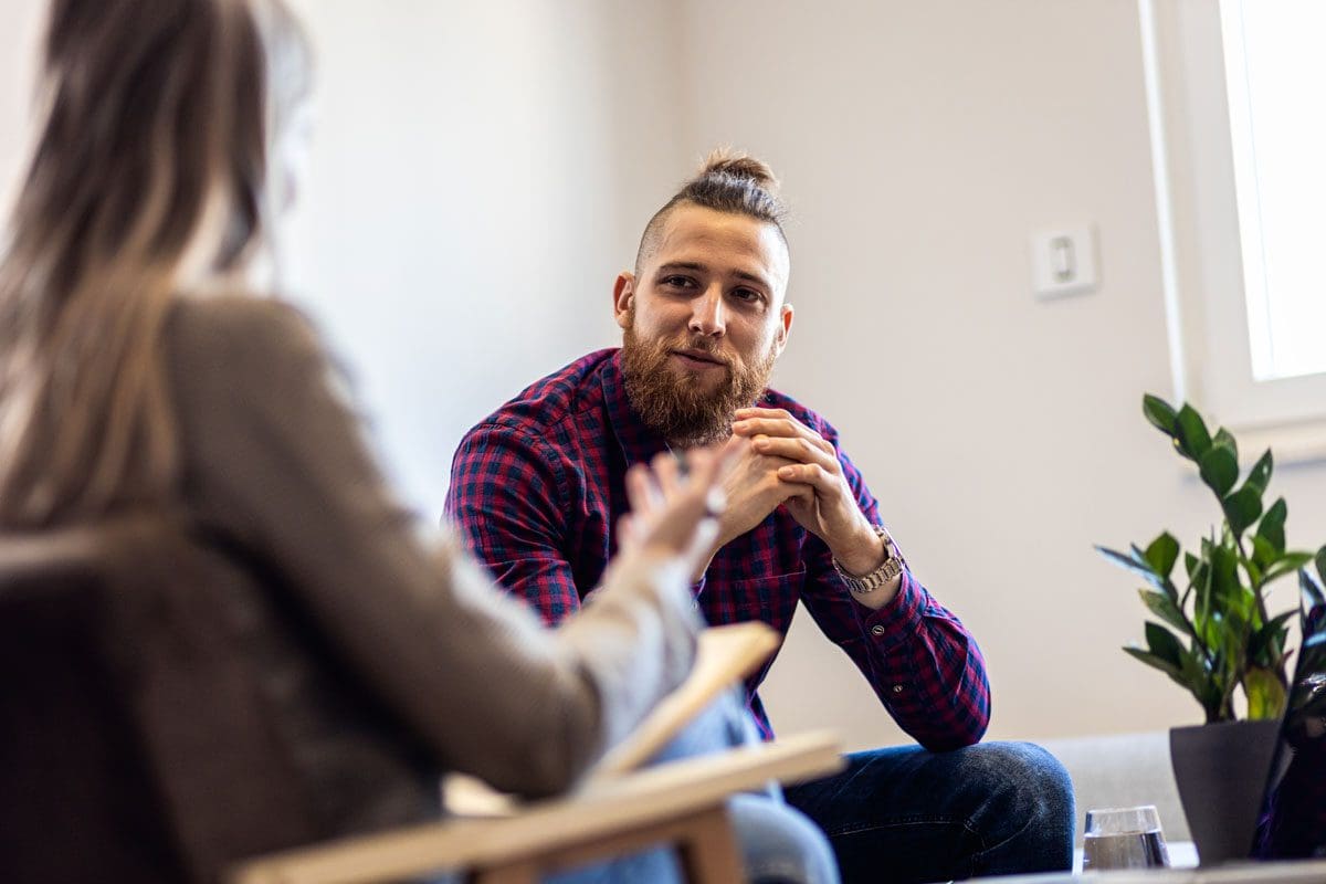 Man with beard, tied-back hair, wearing a red and blue plaid shirt, sitting and talking with a woman in a therapy session.