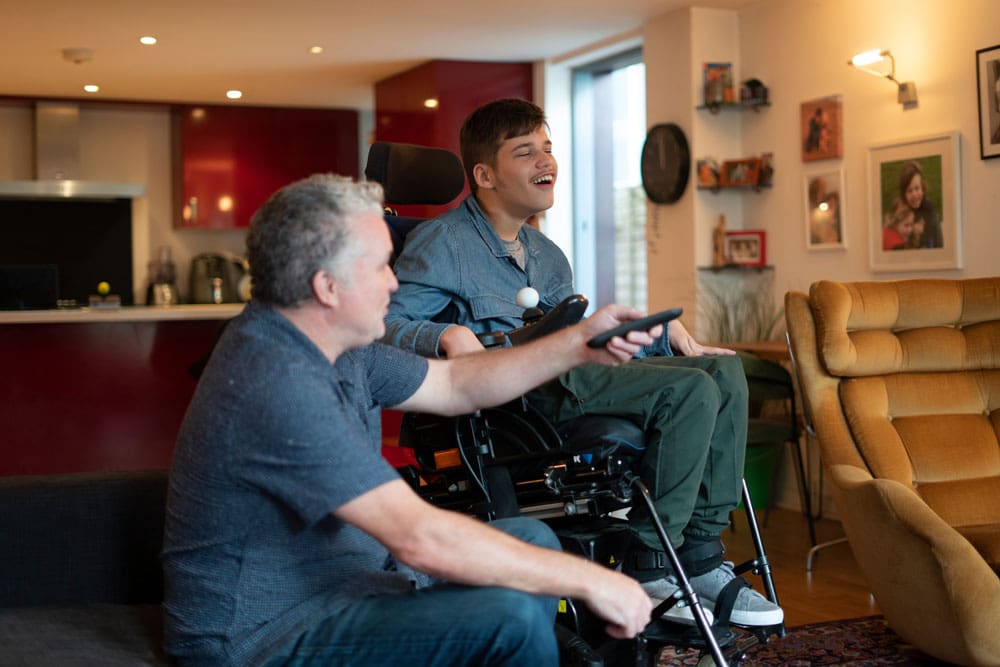 Young man in wheelchair laughing with older man holding a remote.