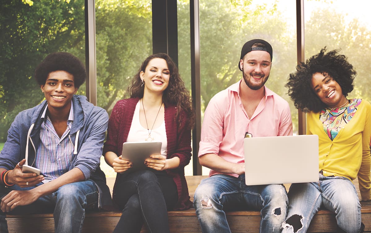 Four young adults sitting and smiling with devices in front of a window.