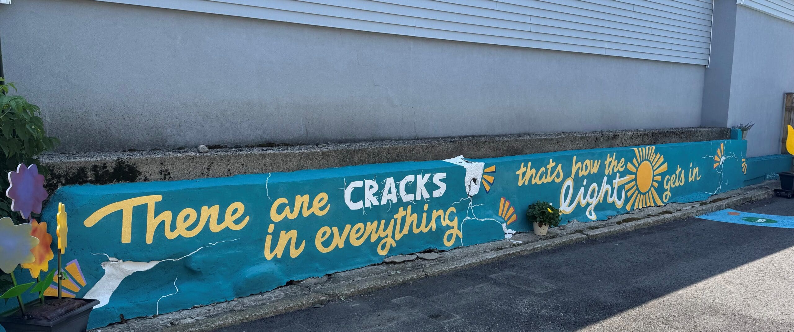 A painted mural on a concrete wall featuring the phrase "There are cracks in everything, that's how the light gets in."