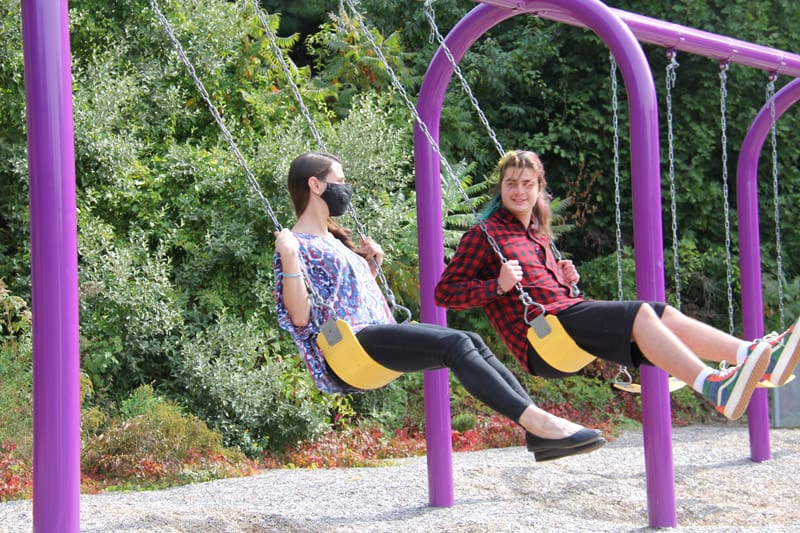 Two teenagers on swings, one wearing a mask, in a park setting.