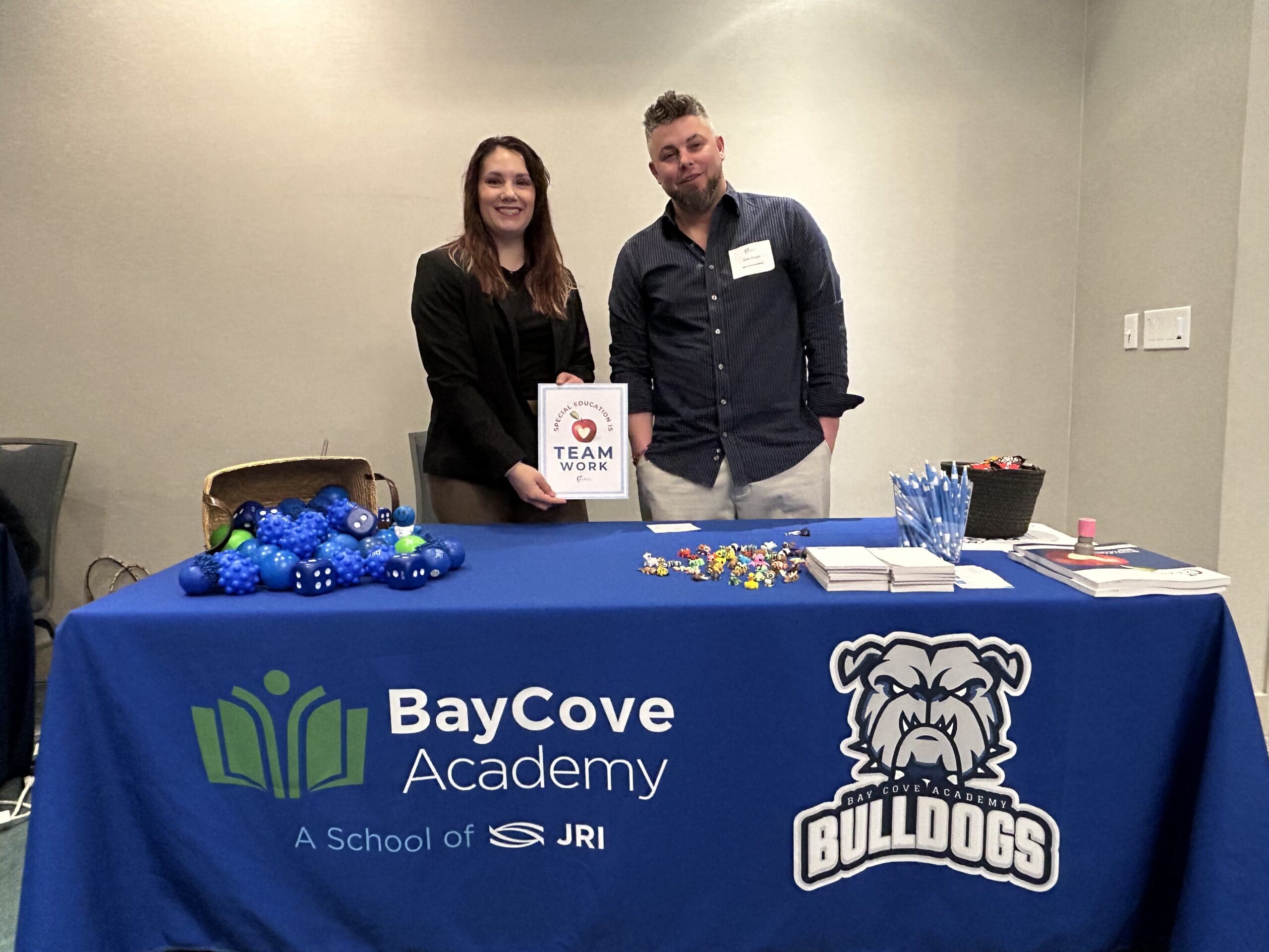Bay Cove Academy staff at an outreach table with branded tablecloth, holding a ‘Team Work’ sign.