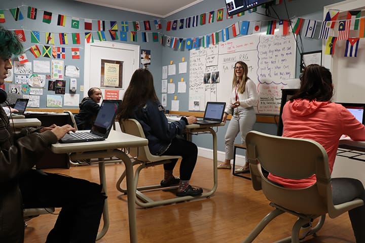 A woman teaching a class to four seated students with laptops in a decorated classroom.