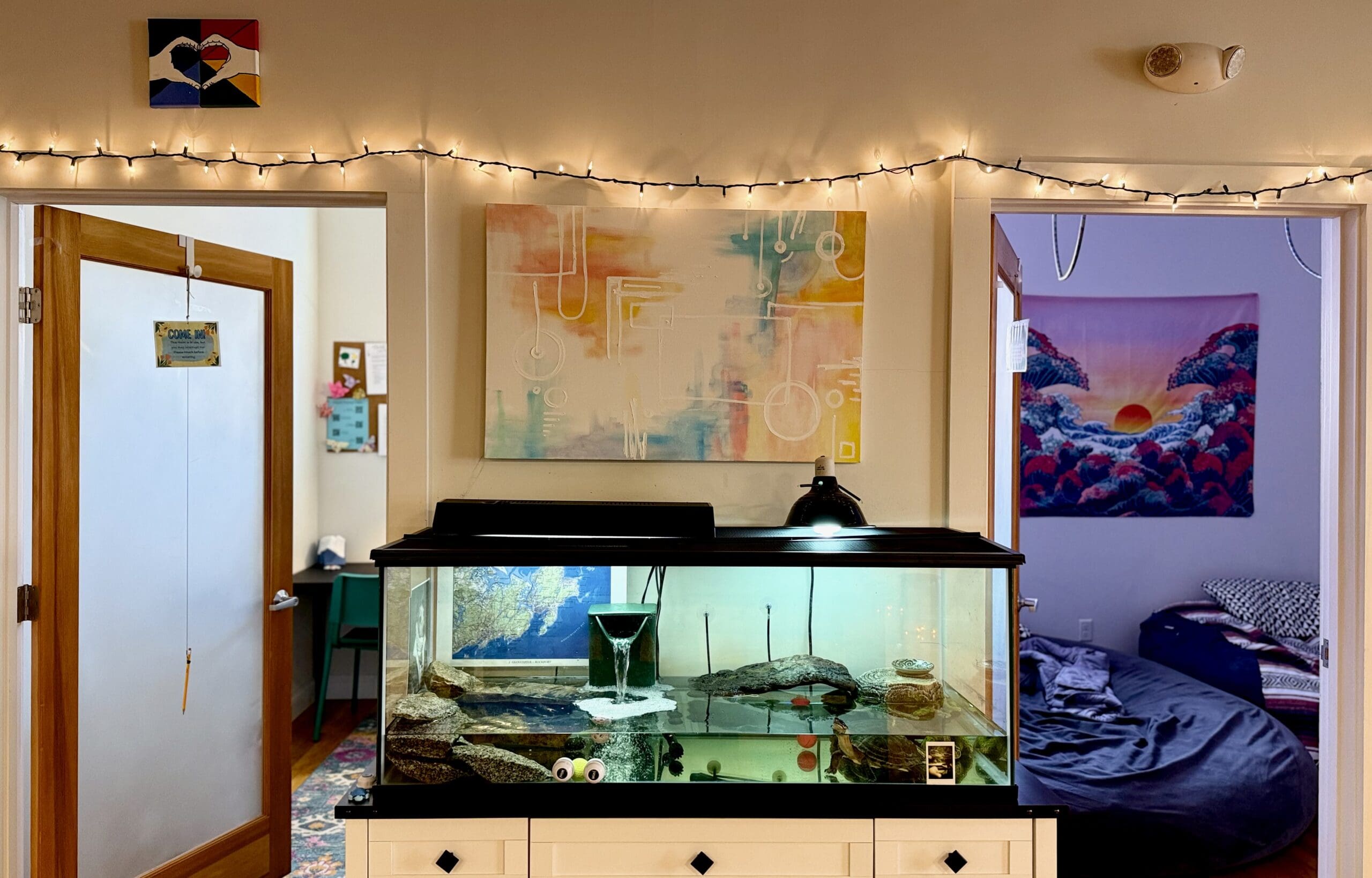 A fish tank on a white stand in a room, with artwork and string lights above, and two doorways.