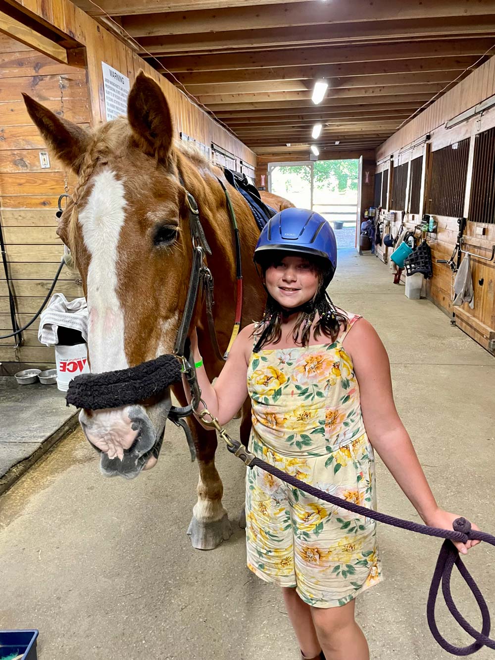 Girl in floral dress and helmet standing next to a brown horse in a stable.