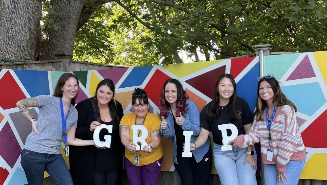 Six people holding letters spelling "GRIP" in front of a colorful mural.