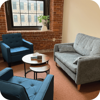 A waiting area with a gray sofa, a blue armchair, a small round table, and a brick wall window.