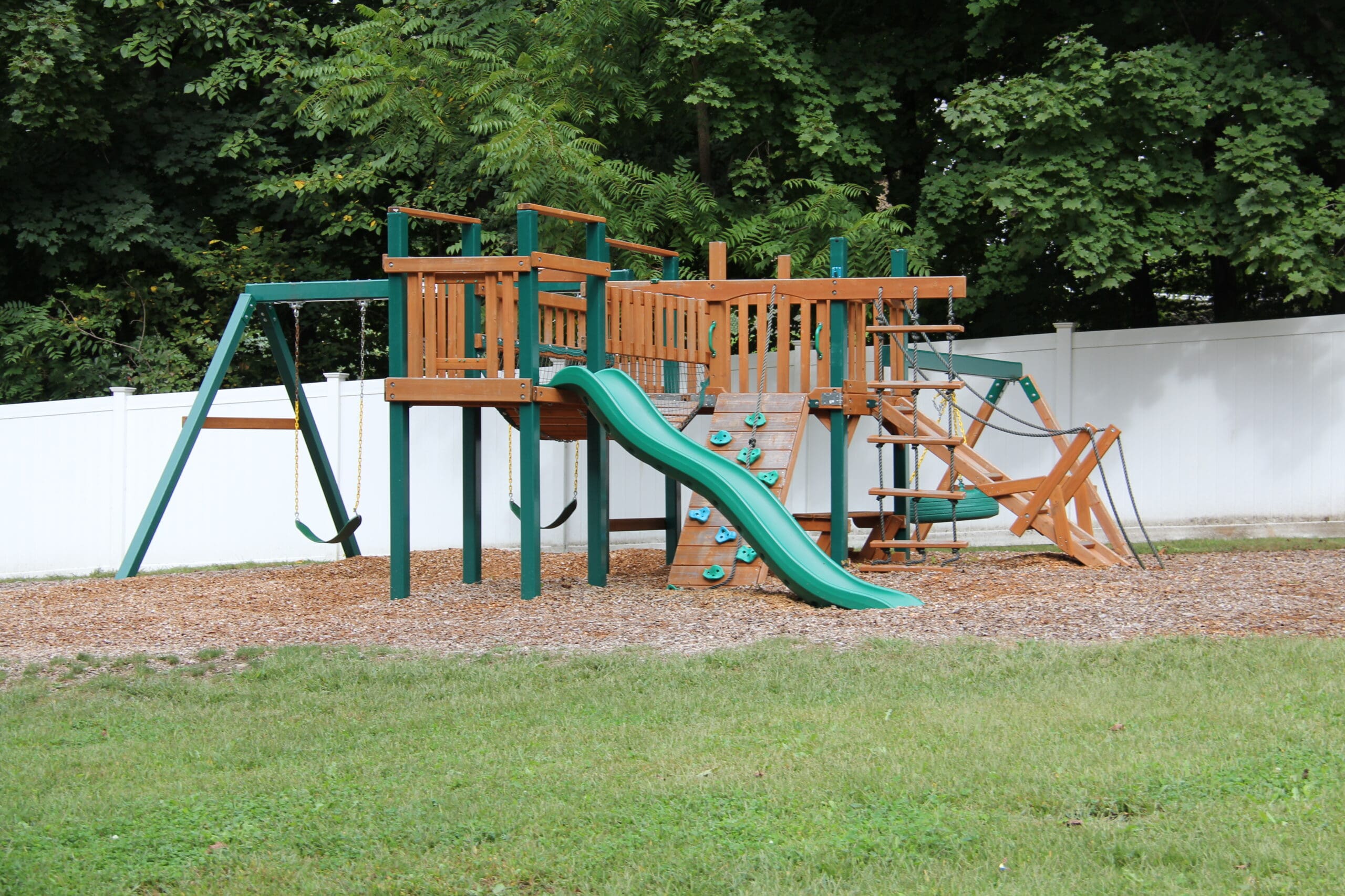 Wooden playground set with slide, swings, and climbing areas, surrounded by grass and trees.